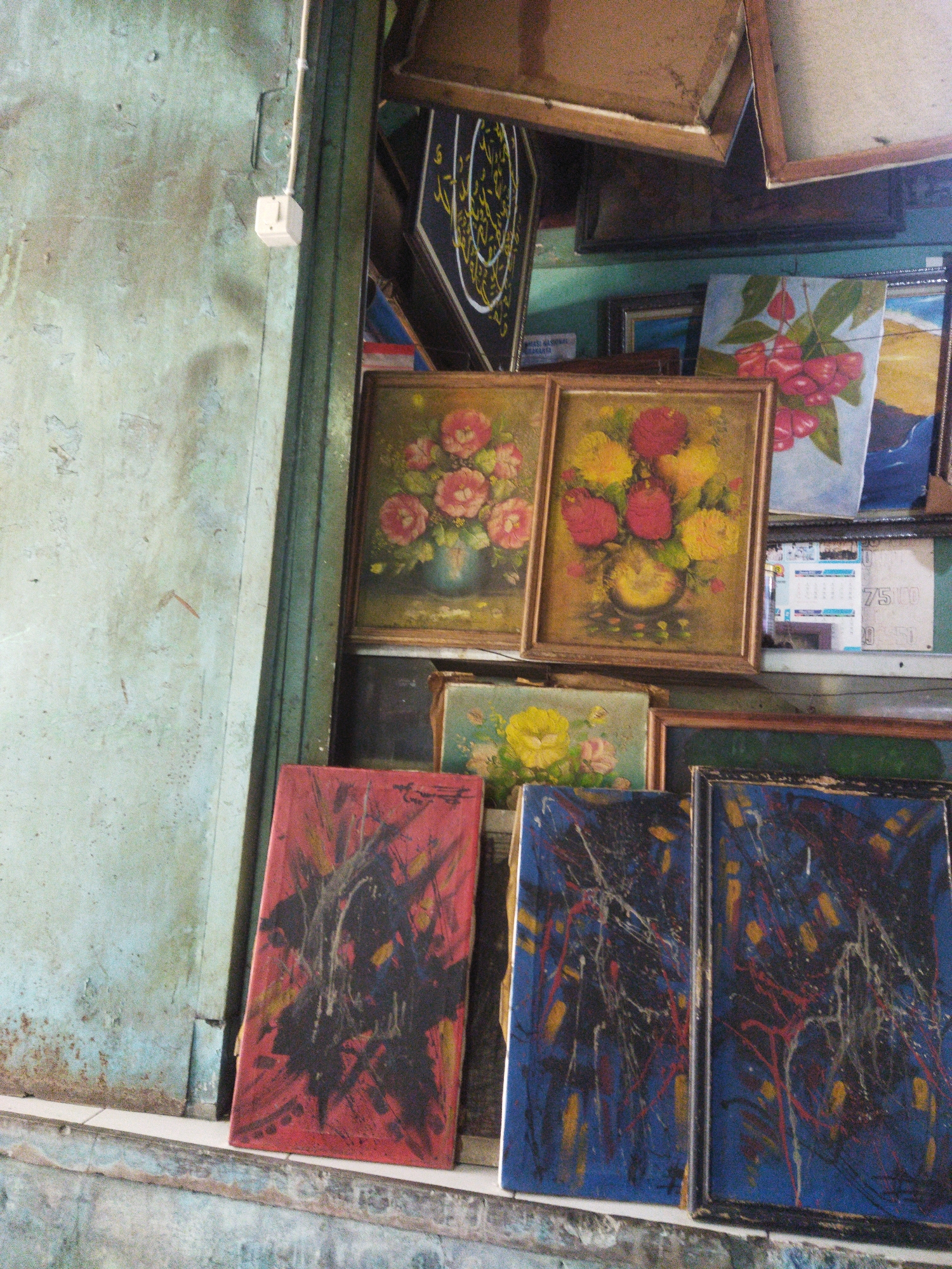 This is a medium shot, eye-level, indoor, full-color photograph of several paintings displayed in what appears to be a shop or gallery. The paintings are of various sizes and styles. In the foreground, on the left, is a painting with a red background and black and yellow abstract designs. To its right are two more abstract paintings with dark blue backgrounds, featuring red, yellow, and white splatters and lines. Behind these, and to the left, are two framed floral still life paintings, one depicting pink flowers in a vase and the other featuring yellow and red flowers. Further back, to the right of the floral paintings, is a larger painting of red cherries on a light blue background. Above these are more framed items, including one with Arabic calligraphy. The background wall is a weathered, light green, textured surface, with a white electrical outlet visible on the left. The shop appears somewhat cluttered, with stacked items and a rough wooden ceiling visible at the top. The overall impression is of a display of artwork in a somewhat aged or rustic setting, likely in the city of Surakarta, Indonesia, given the context provided. There are no people visible in the image.