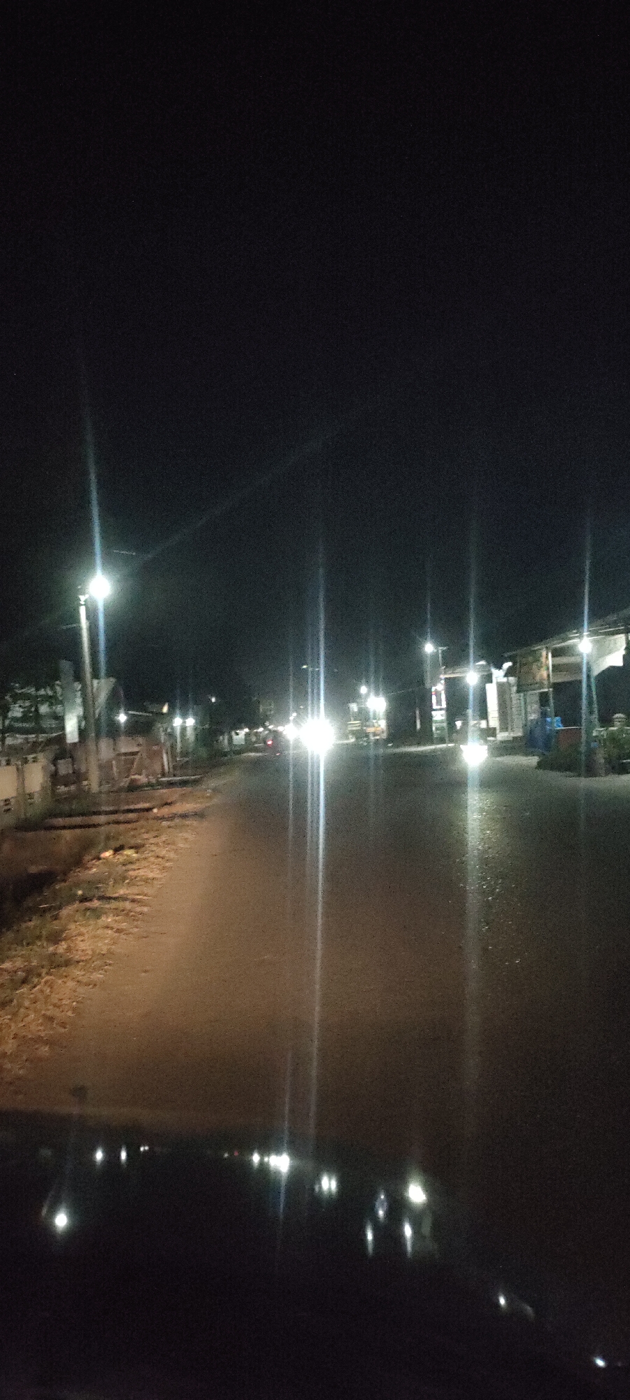 A nighttime street scene in Blangcot Tunong, Indonesia. The image appears to have been taken from a vehicle looking down a road. Streetlights illuminate the roadway, with some buildings and foliage visible on either side. Headlights from distant vehicles are also visible. The foreground is dark, likely the dashboard of the vehicle.