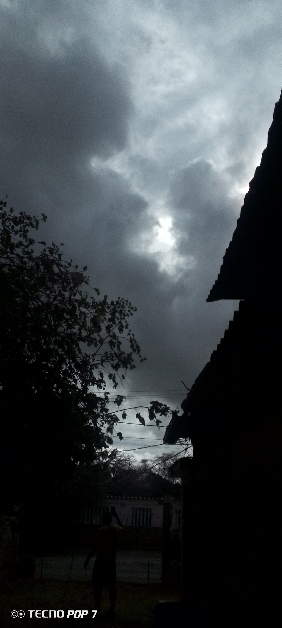 The image depicts an outdoor scene under a heavily clouded sky, captured with a "TECNO POP 7" device as indicated by the watermark at the bottom left. The sky, occupying the upper two-thirds of the frame, is dominated by dark grey and white clouds, with a bright, irregular patch of light, likely the sun, visible through a break in the clouds in the upper-middle right.
In the left foreground, a dense, dark silhouette of a tree or large bush extends from the top left corner downwards, with visible branches and leaves. Below this foliage, electrical wires or utility cables are strung horizontally across the midground.
On the right side of the frame, a distinct, dark silhouette of a corrugated or tiled roof with a steep pitch is prominent, extending from the top to the lower right corner, indicating a nearby building.
In the lower-middle portion of the image, an adult male figure is visible in deep silhouette, standing upright and facing away from the viewer or slightly to the left. He is holding an object, possibly a mobile phone or camera, up to his head or face with both hands, suggesting he is taking a photograph or video. He appears to be wearing shorts.
Behind the figure and the lower part of the tree, a light-colored, single-story building with a darker roof and visible windows or shutters is partially discernible, contrasting with the foreground's shadows. Further in the background, more sparse, possibly bare-branched trees are visible against a lighter horizon. The ground in the foreground is dark and indistinct. The scene is located in Altagracia, Venezuela.