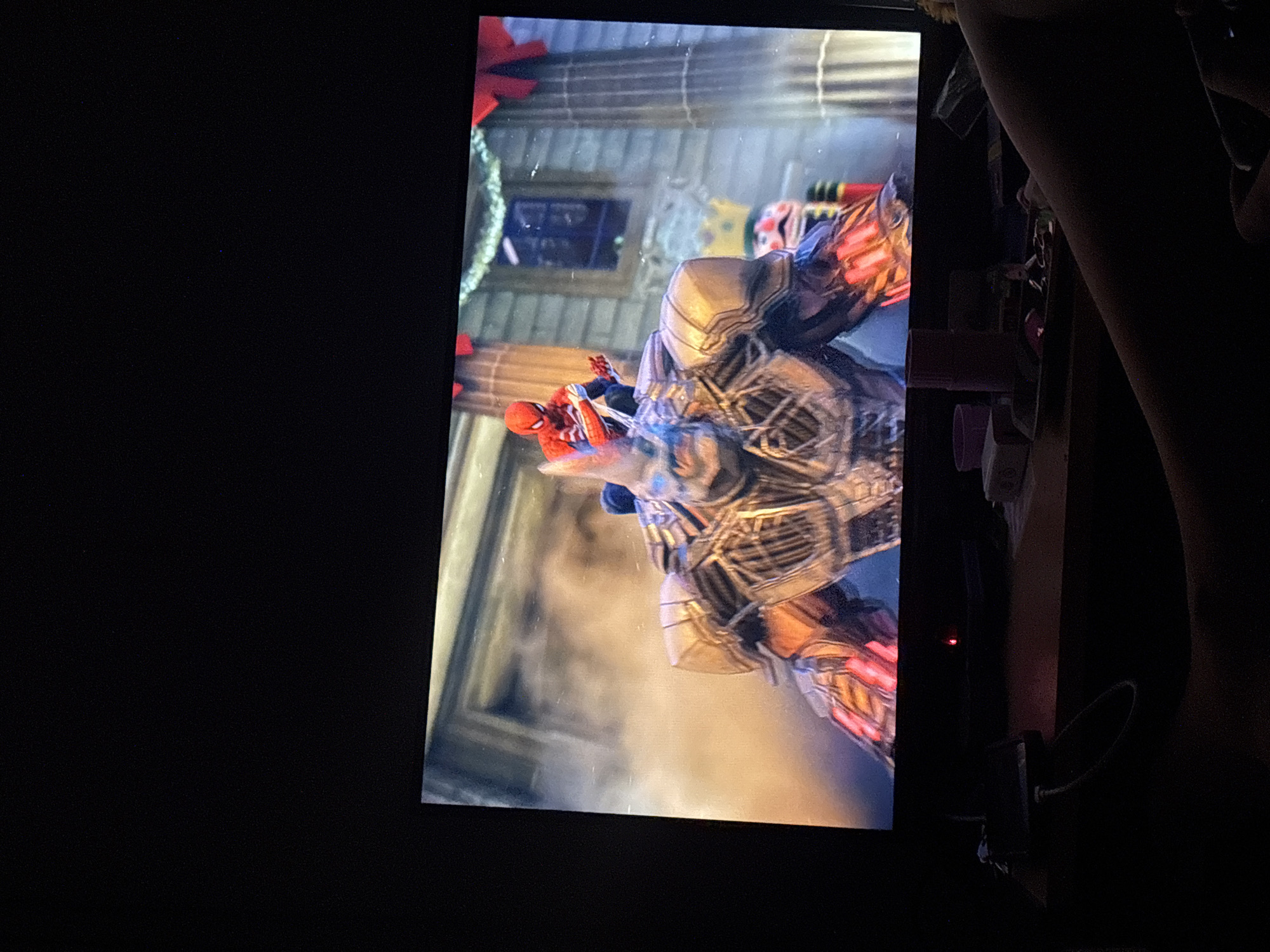 The image displays a screen showing a video game, oriented vertically, within a dimly lit room. The game scene features the superhero Spider-Man in his classic red and blue costume, engaged in an intense battle with a large, armored, mechanical or robotic opponent. This antagonist is primarily metallic, with gold and silver sections, and glowing red elements visible on its forearms or hands, indicating energy or active weaponry. The game's background suggests an urban setting, possibly a city street or building, decorated with red bows and garlands, hinting at a holiday season. Dynamic visual effects surround the characters, emphasizing the action and conflict.
The area to the left of the screen is completely dark, while the right side reveals glimpses of the surrounding environment, also largely obscured by shadow. An arm or hand is visible towards the top right, partially silhouetted. Below this, various indistinct objects, potentially electronic devices or desk items, are visible on a surface. A small red indicator light, possibly from a power source or peripheral, glows faintly on the right edge of the frame, near the bottom of the screen. The screen itself shows minor smudges or dust specks. The overall darkness of the room suggests the image was taken at night or in a low-light setting. There is no legible text visible in the image, nor are there any specific visual cues within the frame to indicate the location as Buenos Aires, Argentina.