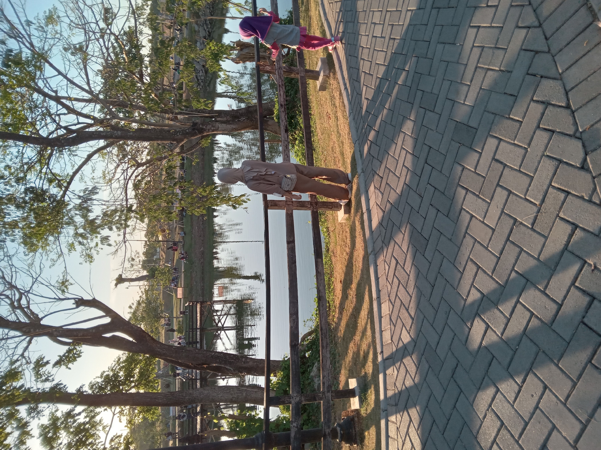 In an outdoor park setting, two individuals are visible near a body of water. In the foreground, a person wearing a brown hijab and matching outfit stands facing away from the camera, with a small crossbody bag. To their right, a child in a purple hijab and pink clothing stands near a wooden fence, looking towards the water. The scene features a paved walkway with herringbone brick patterns, casting long shadows from overhead trees. A dark wooden fence separates the walkway from a grassy area with more trees. Beyond the grass, a calm body of water reflects the sky and trees, with wooden structures and people visible on the far side. The environment appears to be a park or recreational area with a serene atmosphere.