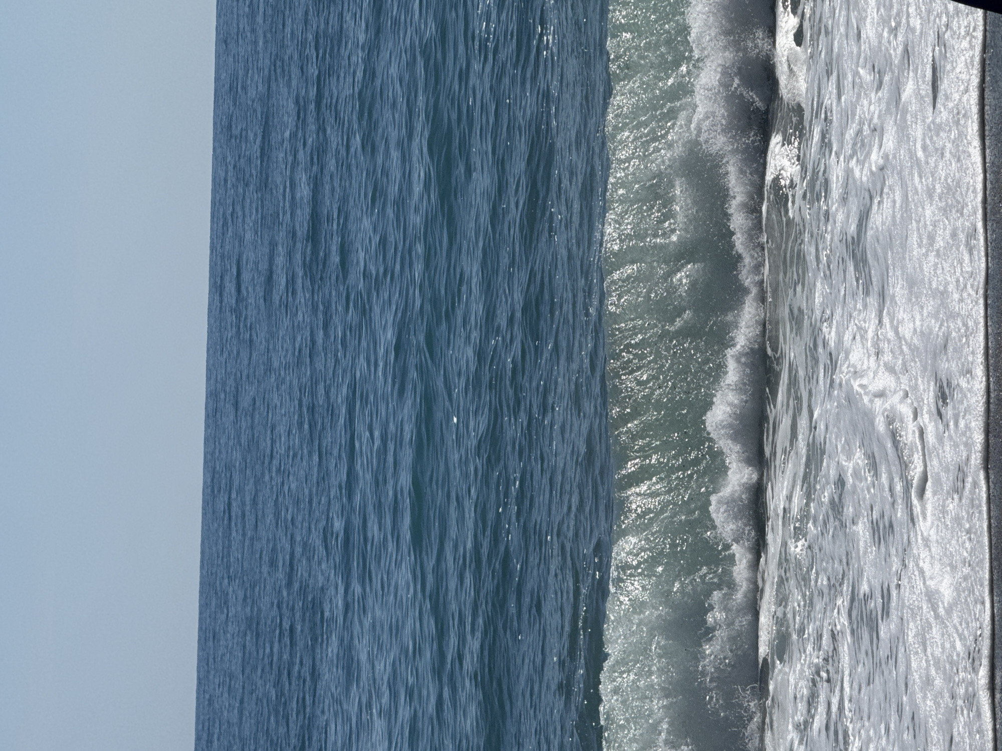 The image shows an expanse of water meeting a textured edge, potentially a beach or artificial barrier. The water transitions from a calm, dark blue on the left to a foamy white near the edge. The sky is a pale blue. No people or distinct objects are visible. The location and city are unknown.
