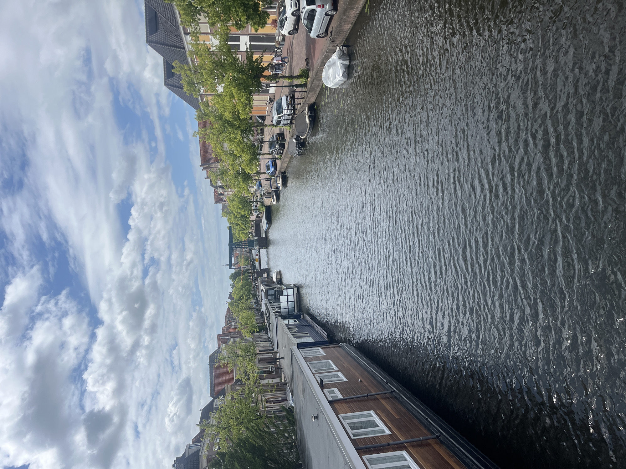 The image shows a canal in a city, likely in the Netherlands given the style of the houses and the canal boats. On the left side of the canal are several houseboats docked side-by-side. The first houseboat is a long, wooden structure with several white-framed windows. Next to it is a darker houseboat with a similar design. Further down the canal, more houseboats are visible.
The right side of the canal features a street lined with trees and buildings. Several cars are parked along the street, and some people are walking on the sidewalk. The buildings are typical of European architecture, with pitched roofs and multi-story designs. A bridge can be seen in the distance spanning the canal. The sky is partly cloudy, with a mix of blue sky and white clouds. The water in the canal is dark and slightly rippled. The overall scene depicts a calm urban environment with a focus on the canal and the life around it.