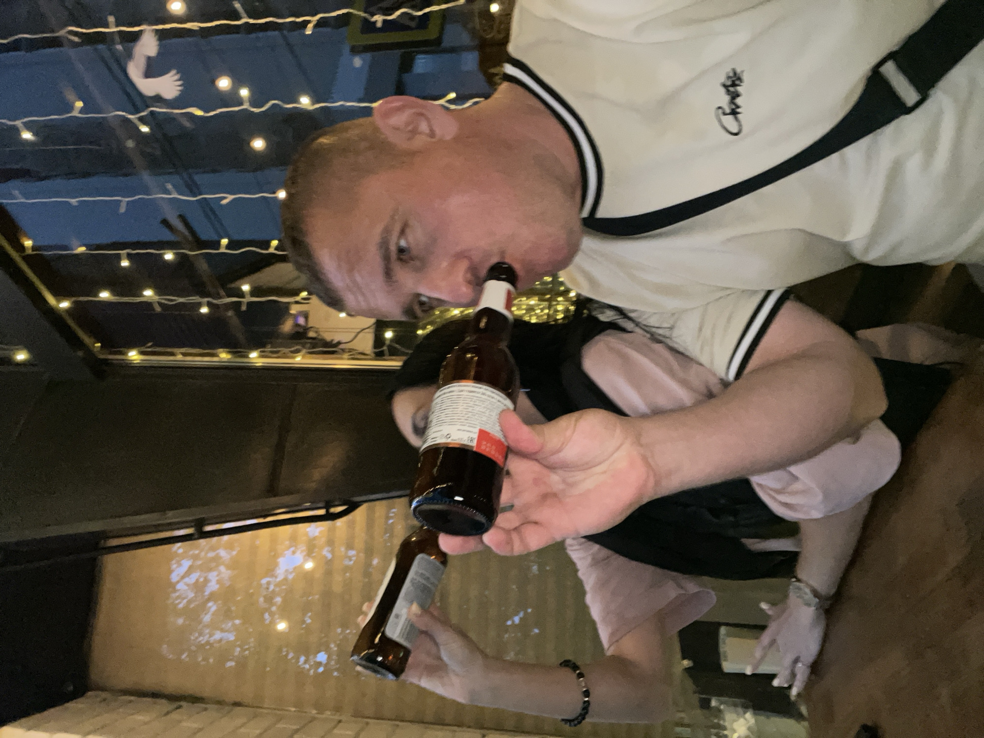 A man is drinking from a brown beer bottle. The photo appears to have been taken at an outdoor bar in the city of Sochi, Russia. String lights and a paper dove hang above the bar. The man wears a white t-shirt with black trim and is holding another beer bottle. A person wearing a pink t-shirt and bracelet stands adjacent to him.