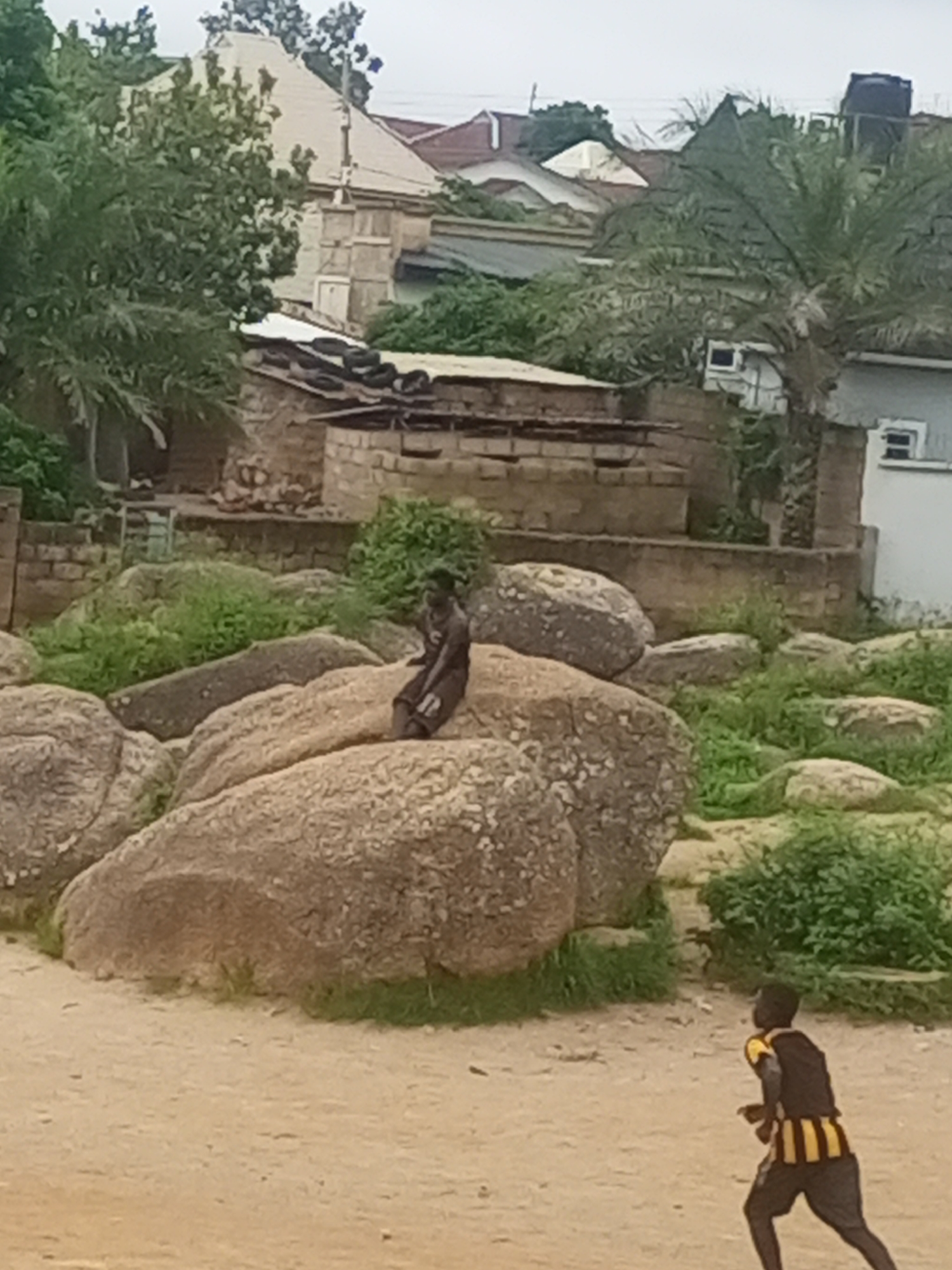 A medium shot shows two young males in an outdoor, somewhat unkempt environment, with rocky outcrops and some vegetation. In the mid-ground, a young Black male is seated on a large, smooth boulder, facing left and looking downwards. He is wearing dark shorts and a dark t-shirt with a white design on the left thigh. The background reveals a mix of structures, including a light-colored, pyramid-shaped roof of a building with stone accents and a dark water tank on a roof. There are also brick structures and some overgrowth. In the foreground, to the right, a second young Black male is in motion, running towards the left of the frame. He is wearing a black vest with yellow stripes and black shorts. The ground is sandy and uneven. The overall impression is one of a casual, informal setting, possibly a residential area in Jos, Nigeria, characterized by natural rock formations integrated with man-made structures.