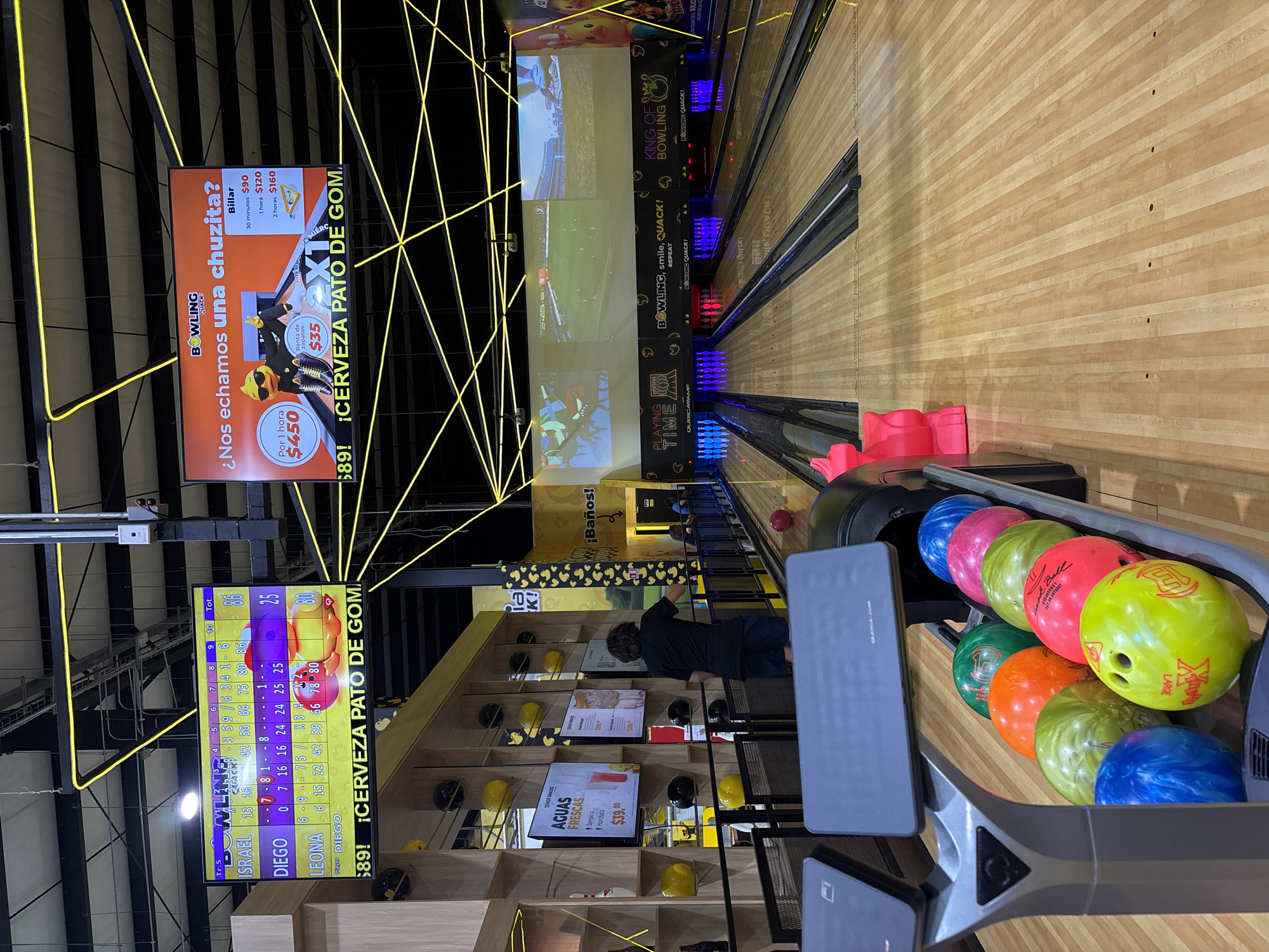 The image depicts an indoor bowling alley at Bolicheria King of Bowling located in Metepec, Mexico. A bowling lane occupies much of the right side of the image with bowling balls in a rack on the foreground to the right. Behind the bowling lane are monitors for multiple lanes that show sports.
On the left of the image are TV screens, each with advertising. The top screen lists prices for playing billiards: 30 minutes for $90, 1 hour for $120, and 2 hours for $160. Shoe rental costs $35, and playing per hour costs $450. The bottom screen displays the ongoing scores of bowlers with the players named Israel, Diego, and Leona.
Behind the screens is a snack bar, with one visible person near the bar. Further back is a sign for bathrooms. Neon yellow light fixtures are arranged in geometric patterns on the ceiling.