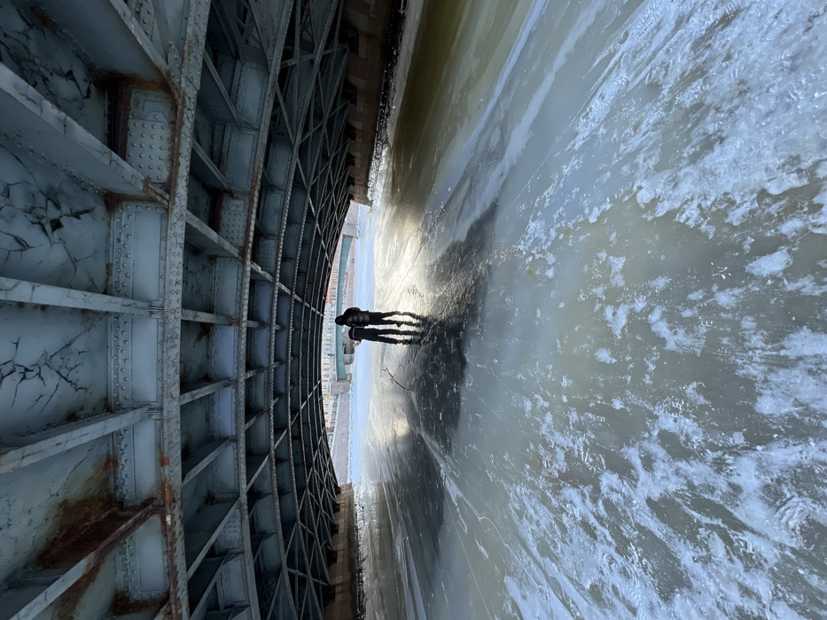 The image depicts a low-angle view from underneath the Pochtamtsky Bridge (Post Office Bridge) spanning the Moyka River in Saint Petersburg, Russia, during winter conditions.
The foreground is dominated by the underside of the Pochtamtsky Bridge, an arched metal truss structure exhibiting a light blue-grey paint finish with extensive rust and areas of flaking, cracked paint. Numerous rivets are visible across the structural beams. The main bridge forms a large, dark arch over the scene.
The Moyka River below is largely frozen, covered by a surface of ice and scattered patches of snow, with visible cracks in the ice. Towards the right side of the frame, a section of the river appears to have thinner ice or open, murky greenish-brown water.
Two human figures, silhouetted against a bright reflection on the ice, stand near the center of the frozen river, directly beneath the main bridge structure. Both individuals are clad in dark, heavy winter coats. One figure appears slightly taller and stands marginally behind the other. Their reflections are visible on the ice surface.
In the background, a second, smaller bridge, identified as the Krasnoflotsky Bridge, spans the river further downstream. Distant buildings line the riverbanks beyond this secondary bridge. The overall lighting suggests an overcast day with a low light source creating strong backlighting and reflections on the ice.