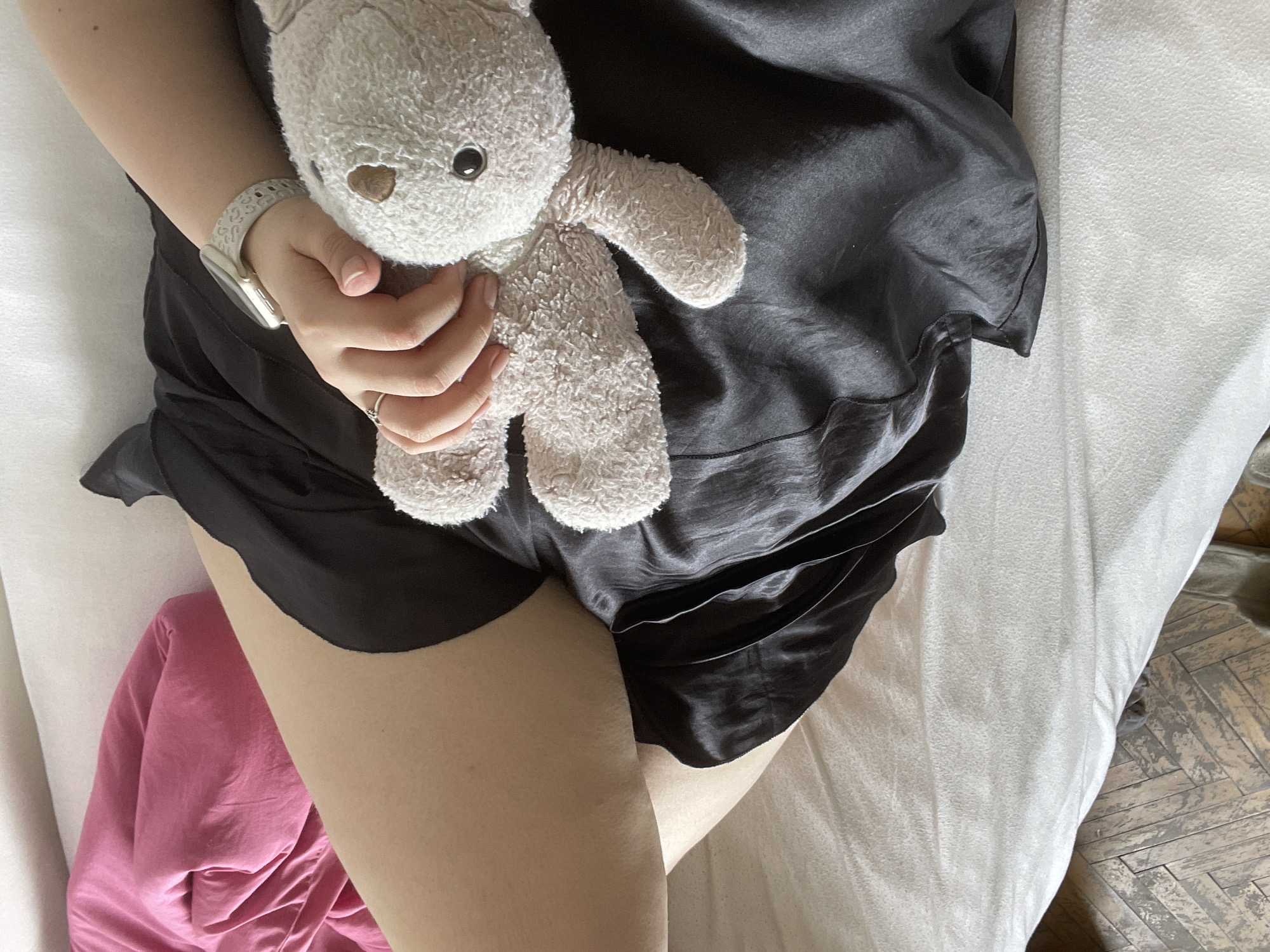 An aerial close-up depicts a person lying on a white bed holding a stuffed animal. The person is wearing a smartwatch on their left wrist and a ring on their left ring finger, and black silk shorts. Pink fabric is visible in the lower-left corner. The location is not discernible from this image, so information about Plovdiv, Bulgaria, cannot be deduced.
