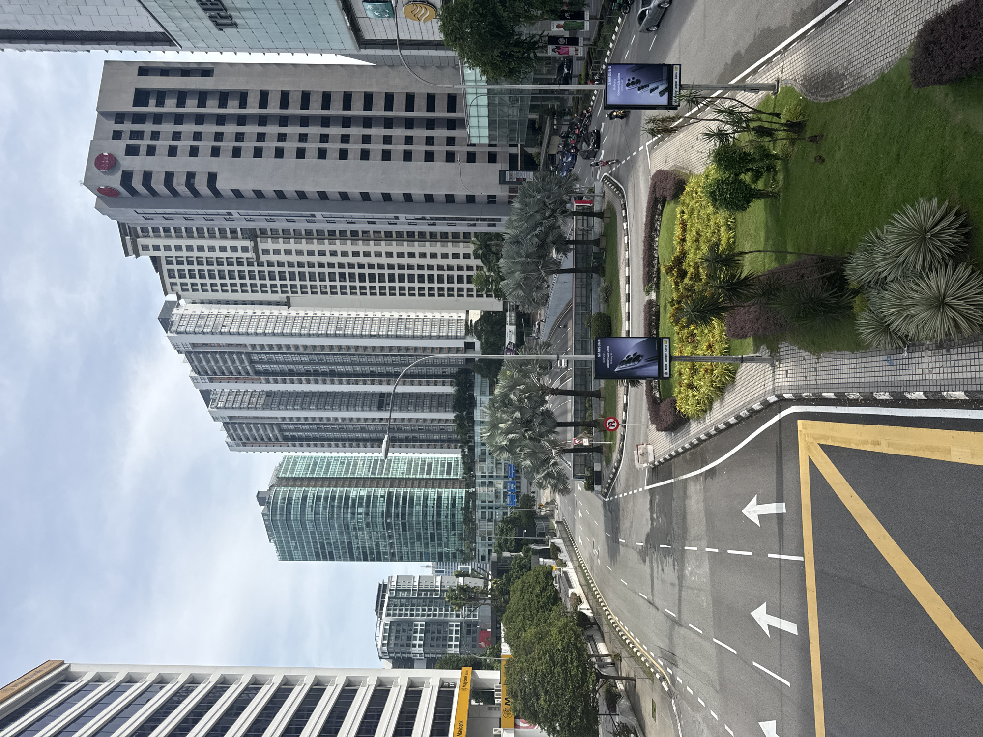 The image captures a high-angle view of a multi-lane urban street in Kuala Lumpur, Malaysia, lined with modern high-rise buildings and lush green landscaping.
**Subjects and Scene:** The foreground features a wide asphalt road with clear white arrows indicating traffic flow (straight and straight/left turn) and dashed lane markings. A large yellow chevron marking is prominent in the immediate foreground. To the right of the road, a landscaped median strip separates the road from a sidewalk, featuring green grass, various shrubs, and several palm trees. On the left, a row of tall, modern buildings dominates the skyline.
**Notable Details and Activity:** The buildings display diverse architectural styles, from glass facades to concrete structures with repetitive window patterns. One building near the top of the frame features a distinct red circular logo with white elements, likely CIMB Bank. Further down the street, another building clearly shows "MAYBANK" and its iconic tiger logo. Several motorcycles are visible, both parked and in motion, alongside a few cars, indicating regular urban traffic. Two large digital billboards prominently display "SAMSUNG Galaxy S23 Ultra" advertisements, with one also bearing the text "A Kuala Lumpur Smart City." Streetlights and a red circular regulatory sign are also visible.
**Time and Weather:** The scene is illuminated by bright, even daylight, suggesting it is daytime, possibly under an overcast or partly cloudy sky, as shadows are soft and diffused. The atmosphere appears calm and typical of a busy city environment.