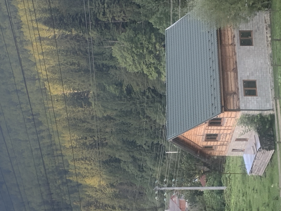 The image displays a two-story residential structure situated on a grassy, uneven slope in what appears to be Tatariv, Ukraine. The primary building features a dark green, corrugated metal roof with visible snow guards. Its upper level is constructed from horizontal light-colored wooden logs or planks, featuring multiple dark-framed windows, with three visible on the main facade and one on the gable end. The lower level is constructed from grey concrete blocks, with four dark-framed windows visible on the main facade. To the immediate left of the house, a stack of light-colored lumber is observed, partially covered by a transparent tarp. A low, wooden fence is partially visible in the lower right foreground.
In the midground, multiple utility poles are present, with numerous black electrical or communication wires extending horizontally across the frame. Portions of other residential buildings, including one with an orange-red roof, are visible to the left of the main structure. The background consists of a dense, steep hillside entirely covered by a mature coniferous forest, with areas of lighter green foliage indicating sunlight exposure. No individuals, animals, actions, or interactions are present in the scene.