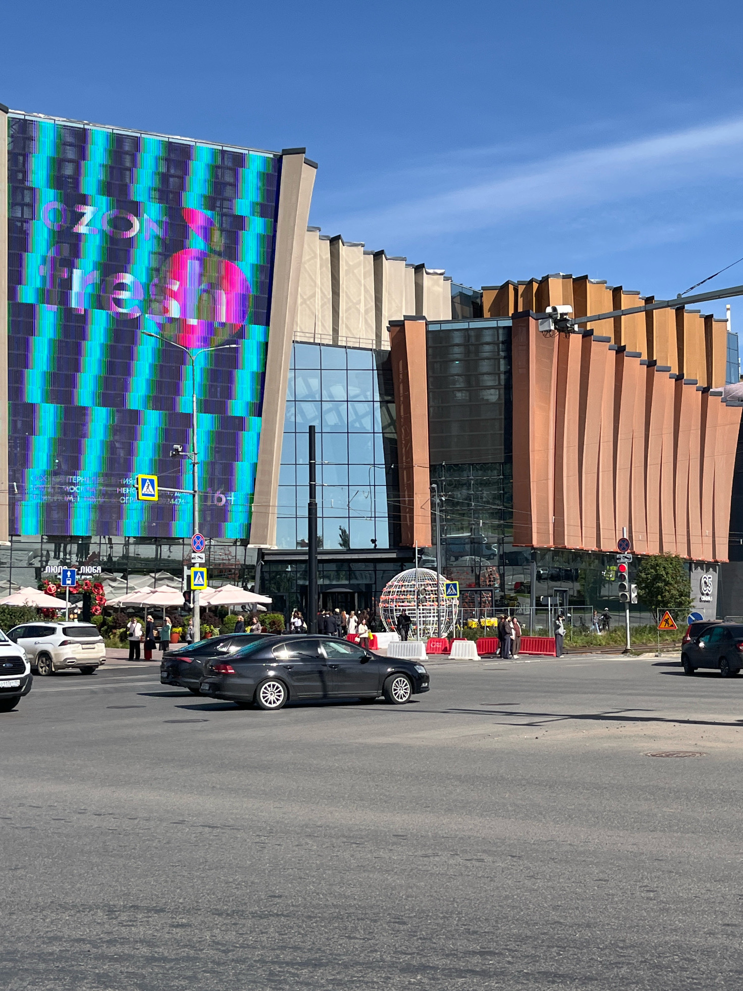 The image depicts a street view in front of a modern building complex in the city of Perm, Russia. A large digital billboard on the left side of the building displays colorful, abstract patterns and text, including "OZON" and possibly "fresh". The building itself features a mix of architectural styles, with a glass facade, beige vertical elements, and reddish-brown paneling.
In the foreground, a street intersection with traffic lights is visible. Several cars are present, including a dark sedan in the left lane and a white SUV further back. Pedestrians are seen on the sidewalk and crossing the street, with one person in a yellow vest standing near a crosswalk sign. A large, spherical light installation is positioned on the sidewalk in front of the building. The scene is set under a clear blue sky on a sunny day.