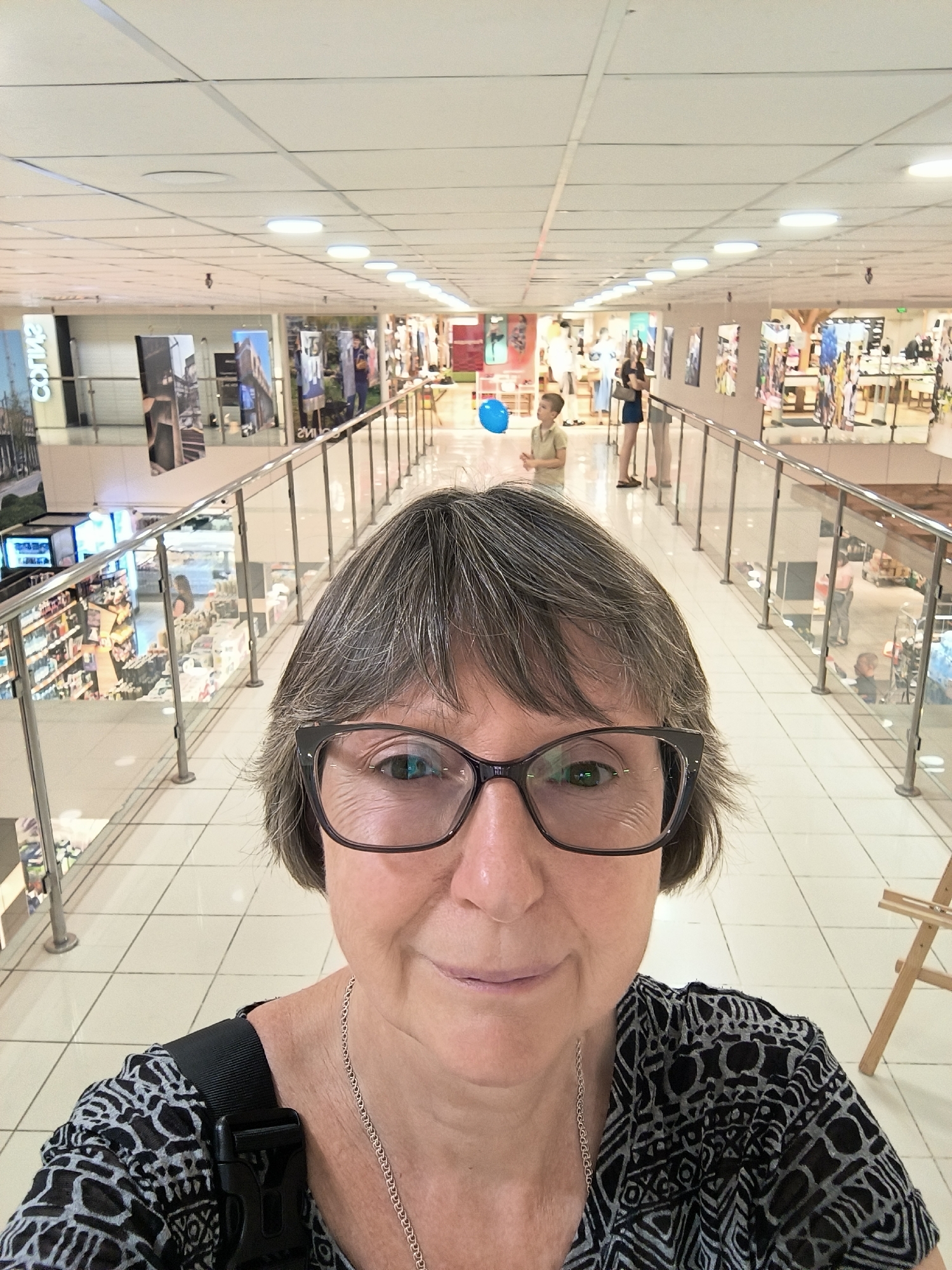 A woman with short gray hair and glasses takes a selfie in a brightly lit indoor space with tiled flooring. She is wearing a black and white patterned shirt and a silver necklace. Behind her, a walkway with glass railings overlooks a lower level with shops. A young boy in a light-colored shirt is visible in the background holding a blue balloon. Several other people are seen in the shops and on the walkway. The environment appears to be a shopping mall or department store. The image does not contain any specific landmarks or contextual clues that would allow it to be definitively located in Mykolaiv, Ukraine.