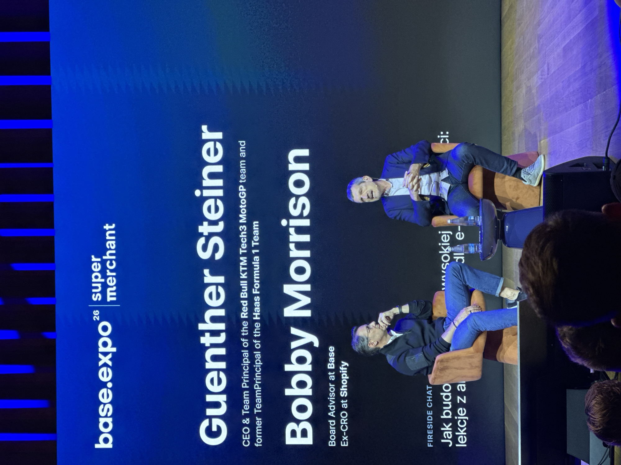 Here is a detailed description of the image:
**Scene**: A photograph of a presentation or discussion event, likely taking place at a conference or exhibition.
**Setting**: The event is related to "base.expo" and "super merchant," suggesting it's a business or technology-focused gathering. The background is a screen displaying the names of two speakers: "Guenther Steiner" and "Bobby Morrison." Guenther Steiner is identified as CEO & Team Principal of the Red Bull KTM Tech3 MotoGP team and former TeamPrincipal of the Haas Formula 1 Team. Bobby Morrison is a Board Advisor at Base and Ex-CRO at Shopify. The title of the session on the screen reads "FIRESIDE CHAT: Jak budo lekcje z a wwwwsokiej dle-ci:".
**People**: Two men are sitting in armchairs on a stage. Their presence aligns with the names displayed on the screen, likely being the individuals named Guenther Steiner and Bobby Morrison, although it cannot be confirmed. There is a glimpse of audience members in the bottom right corner of the shot.
**Objects**: The primary objects are the presentation screen, the chairs, speakers, and possibly water bottles in front of the speakers. The background is partially obscured, but there's indication of a wooden wall and lighting fixtures.
**Actions & Interactions**: The men in the armchairs appear to be engaged in conversation, possibly a discussion or interview. The presence of the audience and stage suggests a public speaking format.
**Environment**: The event is likely taking place in an indoor setting, possibly a conference hall or exhibition center. There are some lighting fixtures present.
**Location**: There are no visual clues that suggest that the location is in the city of Unknown, Unknown.