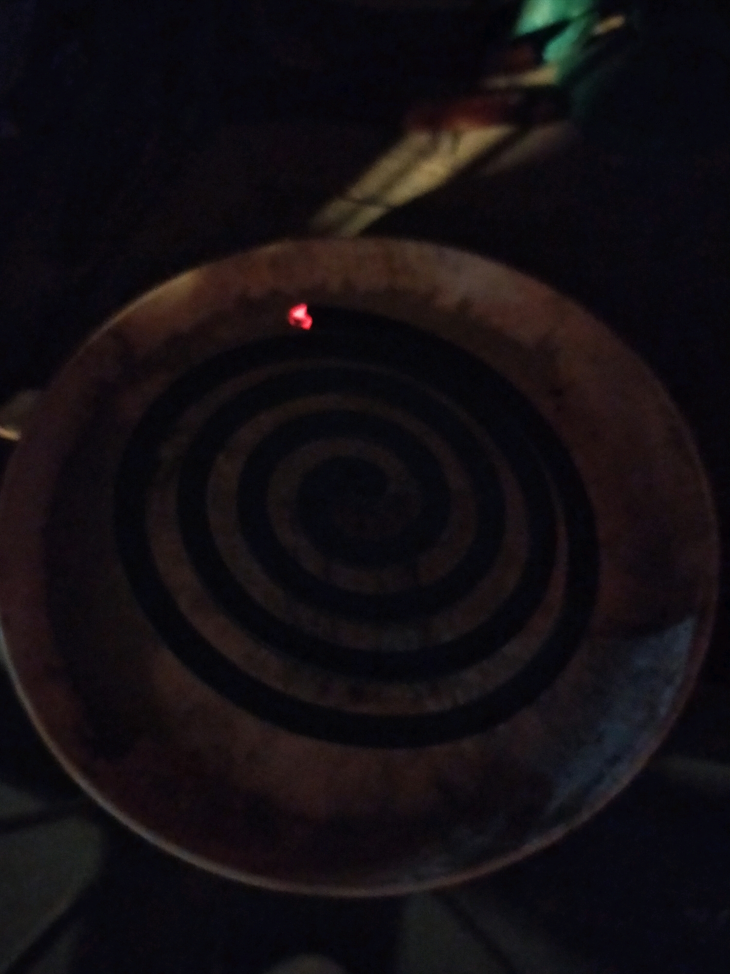 The image displays a close-up, overhead view of a spiral-shaped object, likely an incense coil, with a small glowing red ember at its center, indicating it is lit. The coil is resting on a textured, circular surface, possibly a ceramic dish, with a mottled brown and black pattern. The background is dark and out of focus, with faint hints of green and yellow light, suggesting an indoor setting at night. The scene is devoid of any people, and there are no other discernible objects or activities. The specific location in Surabaya, Indonesia, cannot be determined from the visual information provided in the image.