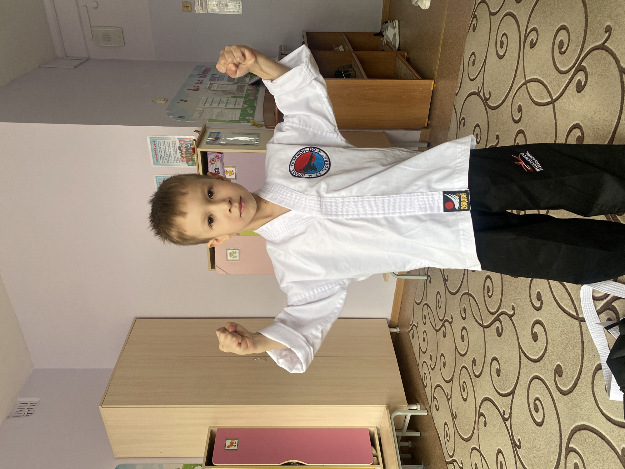 A young boy in a white taekwondo uniform (dobok) with black pants strikes a martial arts pose, holding his fists clenched and ready. He stands indoors, likely in a school or sports club in Chaikovsky, Russia, given the Russian text visible in the room. The boy has a serious, focused expression as he looks directly at the camera.
The setting features light-colored walls and a patterned carpet with swirling brown designs. To the boy's left, a light-colored wooden cabinet or shelving unit is visible. On the wall behind him, several informational posters are displayed. One poster clearly shows the Russian text "ЗАДАЧИ ГОДА" (Tasks of the Year), and another reads "Для вас, робити!", which is likely "Для вас, родители!" (For you, parents!). An air vent is visible on the ceiling.
The boy's uniform features several distinct markings. A circular patch on his left chest displays the words "ТЭКВОН-ДО" (Taekwondo) and "СК УСПЕХ" (SK USPEKH - "Sports Club Success") in Russian. The label on his white belt reads "ZARUBA". His black pants also feature text, partially visible, which reads "TAEKWONDO INTERNATIONAL". Near his feet on the carpet, a white taekwondo belt is casually placed, suggesting he might be either a beginner or has just changed into his uniform. The indoor lighting suggests no specific time of day.