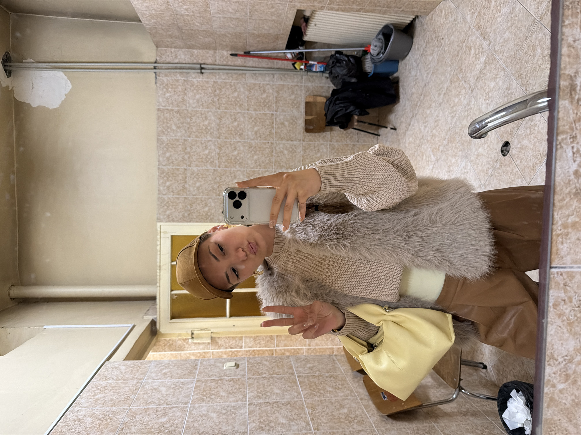 A young woman wearing a brown cap, beige sweater, and a fur-trimmed vest is taking a selfie in what appears to be a bathroom. She is holding an iPhone with a white case. She is making a peace sign with her left hand. A yellow handbag is slung over her shoulder. She is wearing tan-colored pants. The bathroom has beige tiled walls and flooring. A mop, bucket, and other cleaning supplies are visible in the background. A metal bar runs along the wall near the ceiling. A window is visible behind her. The location cannot be determined to be Sofia, Bulgaria based on the provided image.