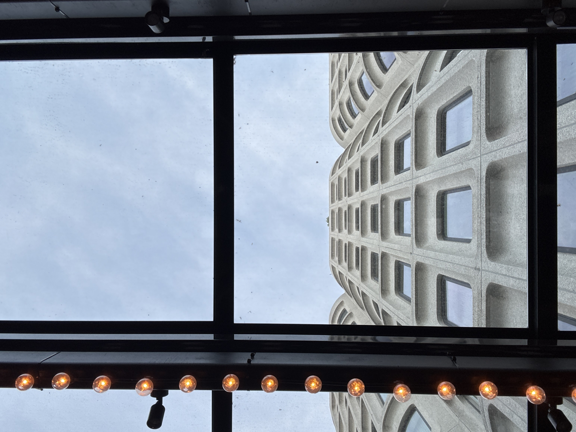 An upward-looking shot through a window features a modern building with a concrete grid facade to the right. The window has a black metal frame, and a string of warm-toned Edison-style light bulbs hang below. The building is viewed against a cloudy sky. Based on the architecture and location, the building is likely 5 Merchant Square, in the city of London, United Kingdom. No people are visible.