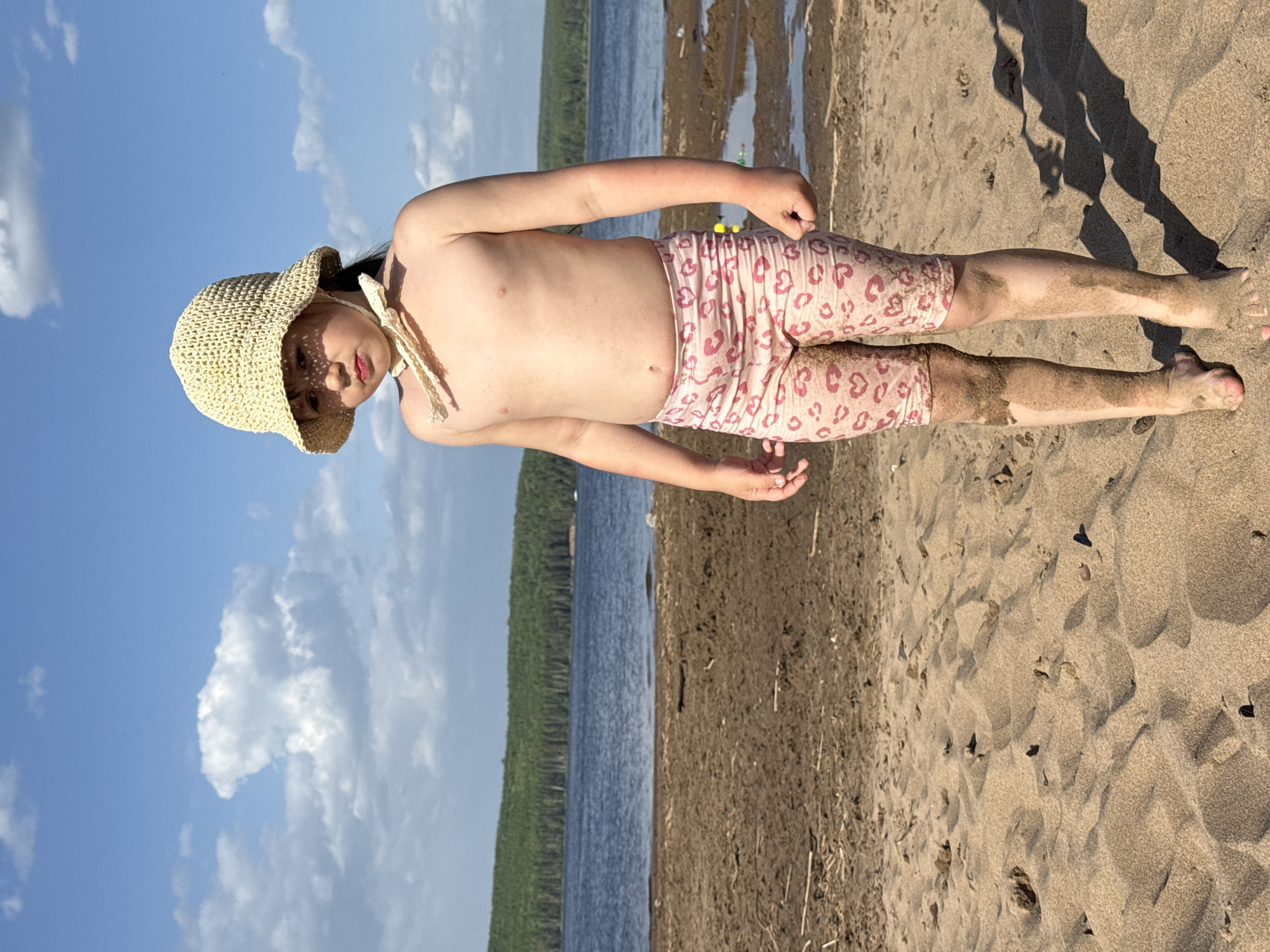 A child wearing a straw hat and pink leopard print shorts stands on a sandy beach. Behind the beach is a body of water with a line of trees visible on the opposite shore. The sky is blue with scattered clouds. The location is likely the city of Chaikovsky, Russia.
