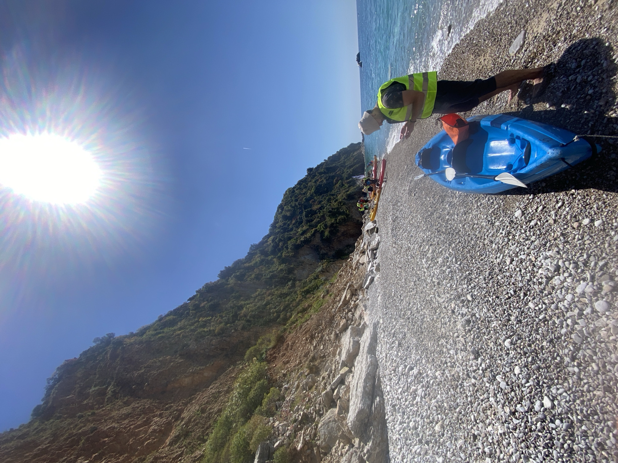 The image shows a pebbled beach on a sunny day, likely in Budva, Montenegro given the coastal environment. In the foreground, a blue kayak with a paddle is beached near the water's edge. A person wearing a high-visibility yellow vest is leaning over, possibly attending to the kayak. In the background, a green, tree-covered mountain rises next to the beach. Further down the beach, other people and colorful kayaks are visible. The sky is clear blue, and a bright sun is visible in the upper left corner. The tide appears to be coming in, as the edge of the beach is wet.