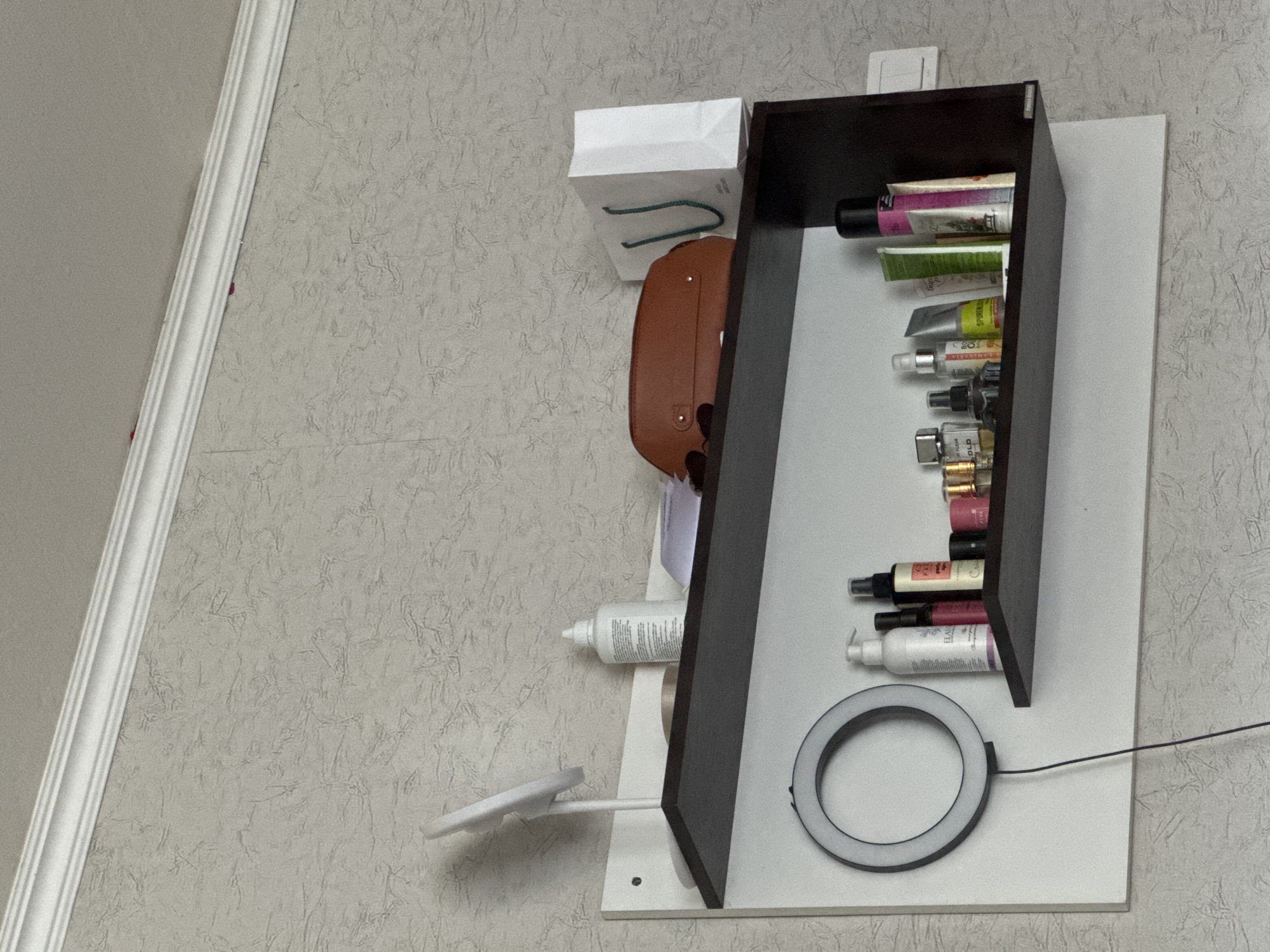 A shelf unit is mounted on a textured wallpapered wall. The shelf unit consists of a white back panel and a dark brown shelf. On the top shelf, there is a white bottle, a brown pouch, and a white paper bag. On the lower shelf, there is a ring light, several bottles of cosmetics and perfumes, and some tubes. The scene is indoors, likely in a domestic setting. The specific location in the city of Яромінскі сельскі Савет, Belarus, cannot be determined from the image alone.