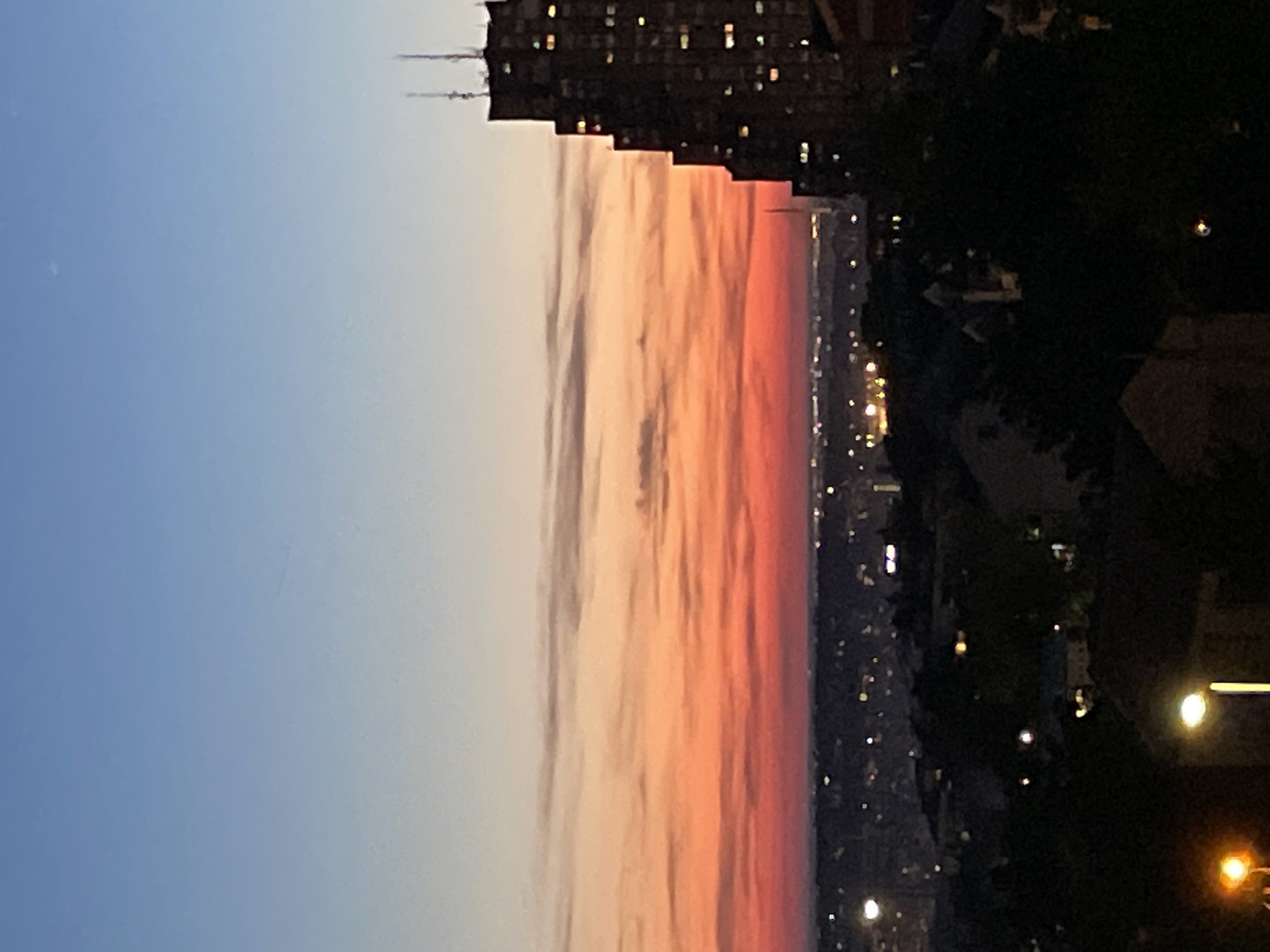 The image depicts a sunset in Belgrade, Serbia. The sky is a gradient of blue to orange and red. A tall, modern building is silhouetted against the sky. Below, the city lights are visible, along with trees and other buildings.
