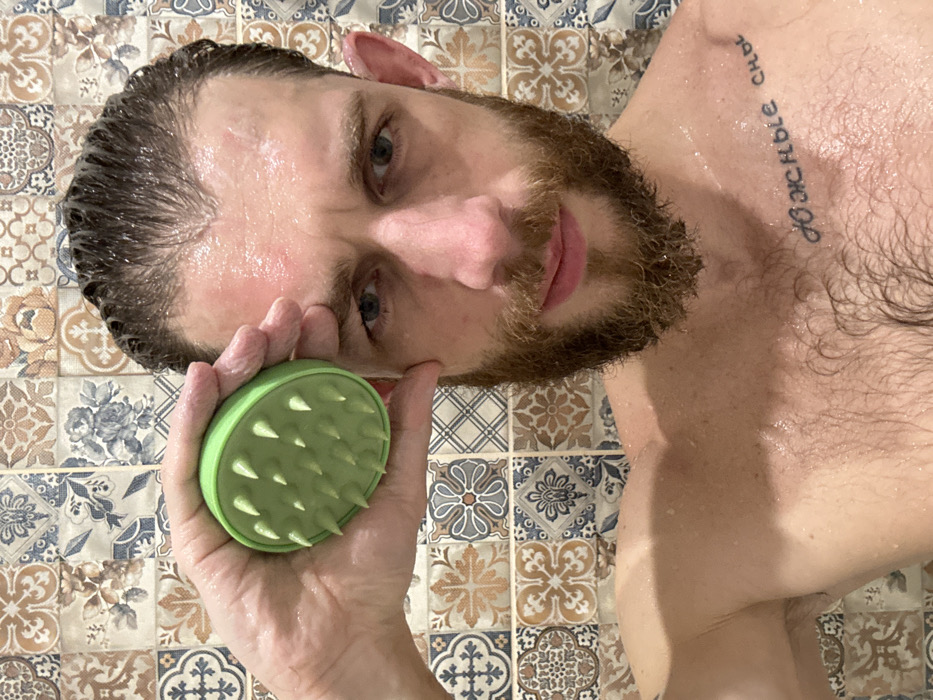 A wet-haired man with a beard and tattoo is holding a green scalp massager with soft rubber bristles. His head is tilted back, and the massager is near his forehead. The background is tiled with a mix of geometric and floral patterns in light browns and blues. He appears to be in a shower, with water visible on his skin.