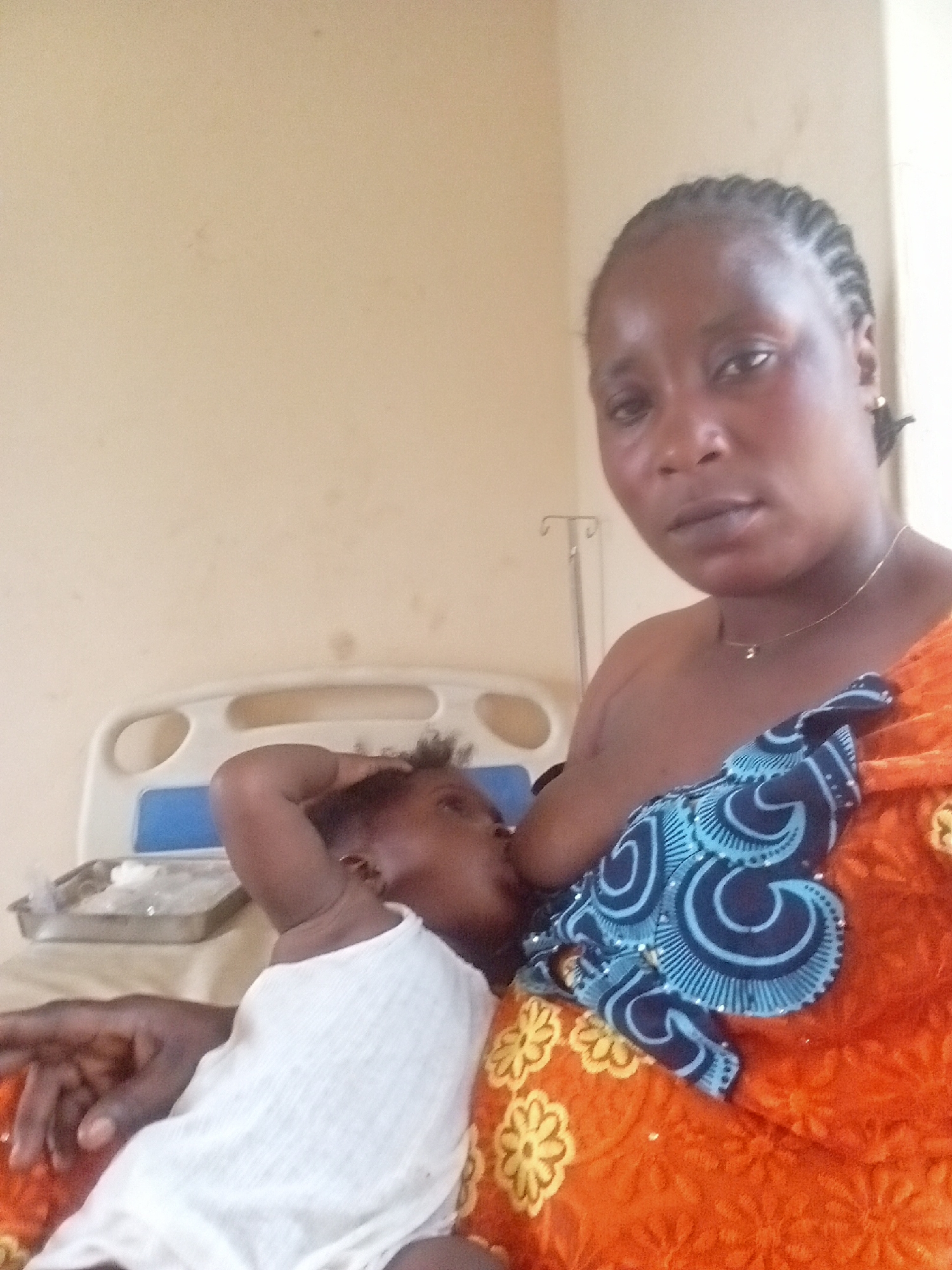 Akwanga, Nigeria: An African woman is breastfeeding her baby while sitting on a hospital bed. The woman has dark skin and braided hair. She wears a gold necklace and is draped in an orange cloth with yellow flower designs. A blue patterned cloth is also present. The baby is fair-skinned and wearing a white top. An IV pole is visible in the background near the cream-colored wall. A metal tray is on the bed, beside the woman and child.