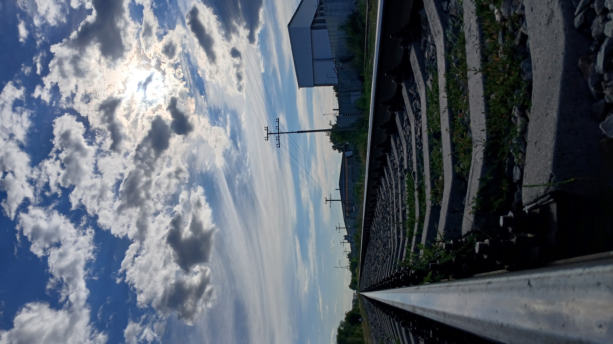 A low-angle shot looks down a set of railroad tracks receding into the distance under a cloudy sky. The sun peeks through breaks in the clouds, casting a bright glow. The tracks are made of metal rails set on concrete sleepers. Grass and small stones are visible between the sleepers. In the background, there are power lines and poles, as well as industrial buildings. The scene is located in the city of Oral, Kazakhstan.
