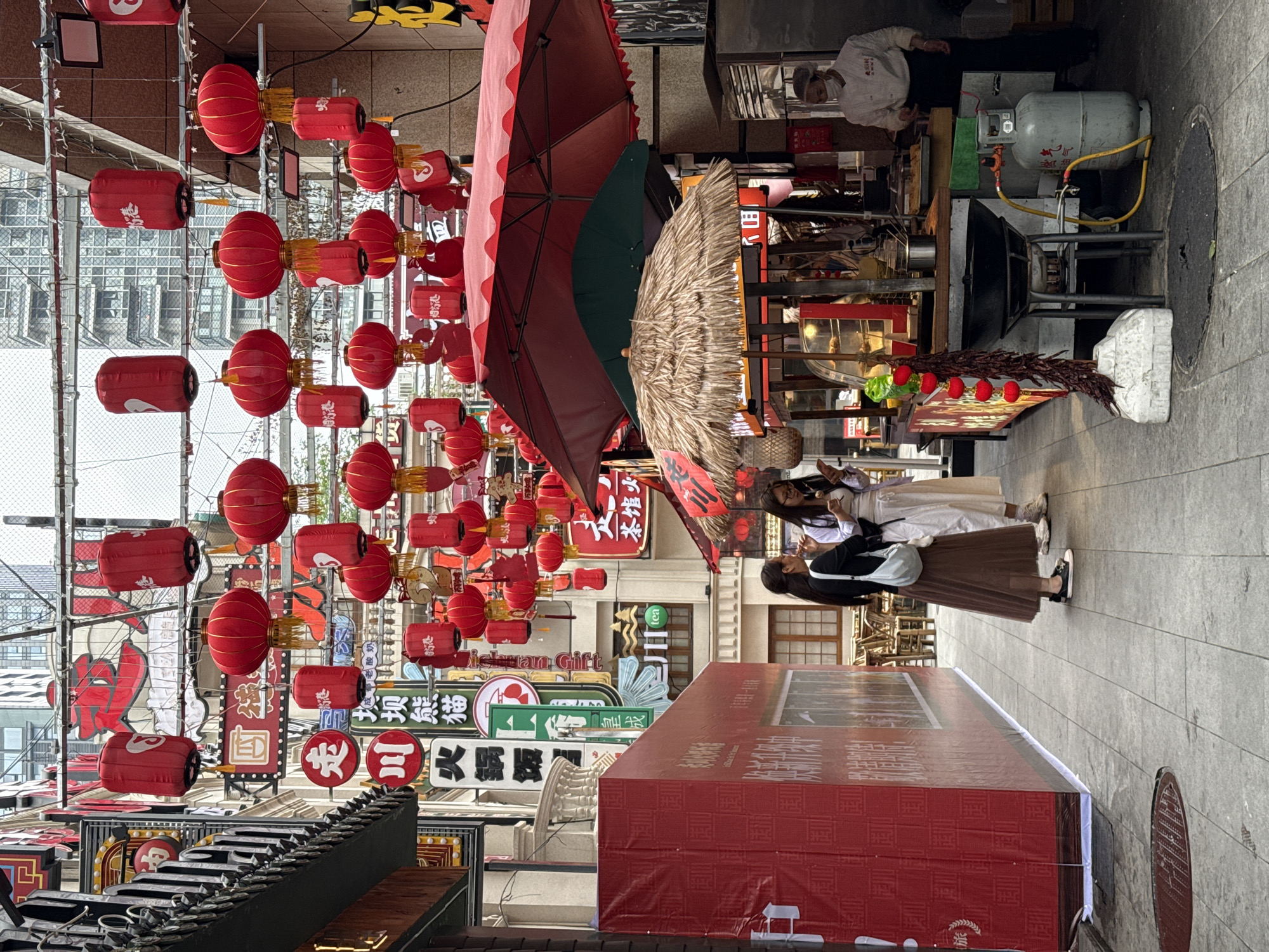 This image captures a vibrant street scene, likely in the Jinjiang District of China, characterized by a dense array of cultural elements and commercial activity.
The foreground is dominated by red lanterns suspended overhead, with various Asian-themed signs in the background. A person wearing a white chef’s coat is seen inside one of the shops that line the street. The street is cluttered with commercial displays, including an outdoor cooking setup involving a wok and a propane tank.
Two women in business attire and wearing masks walk towards the street.