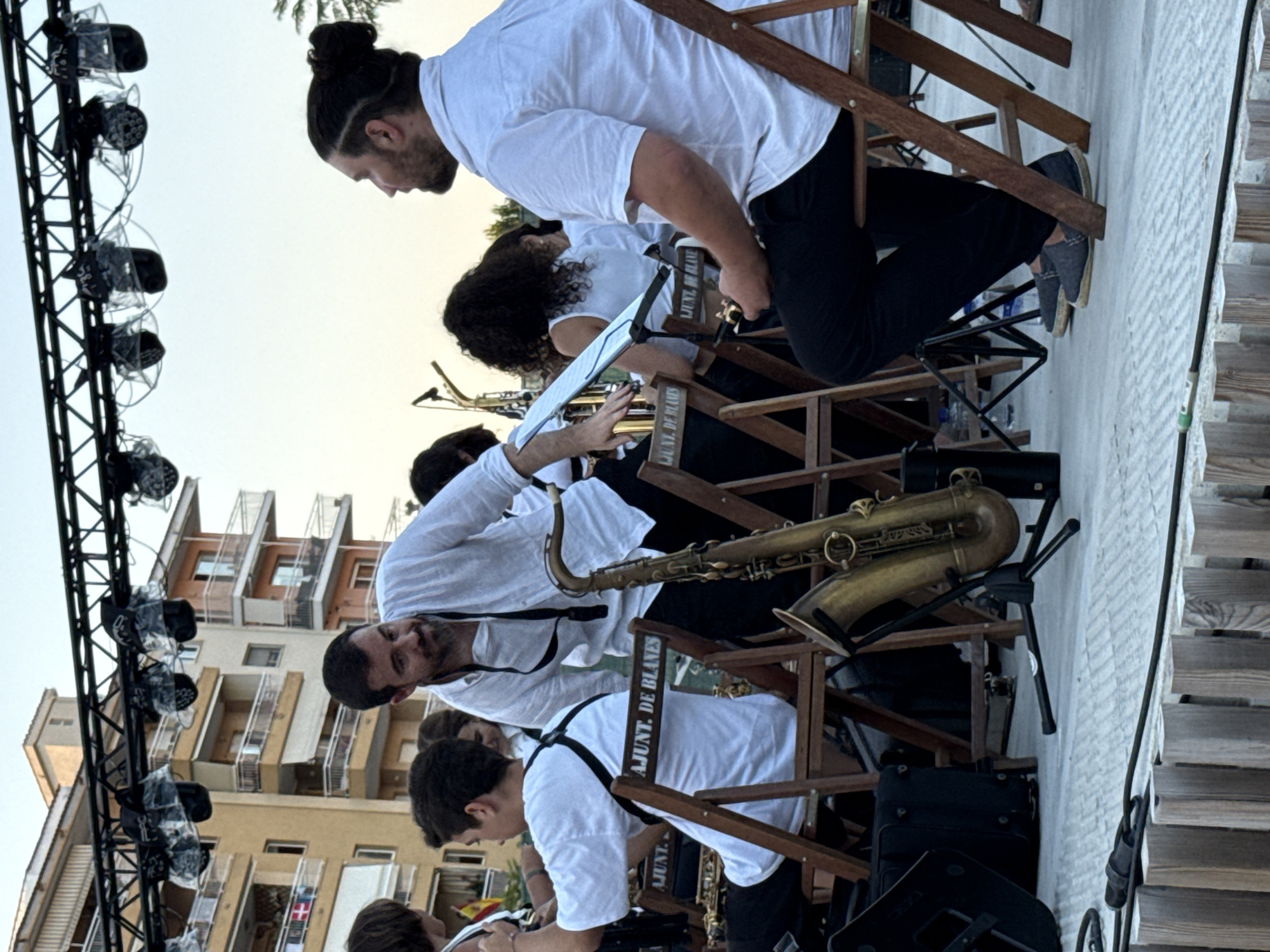 The image depicts an outdoor performance scene, likely in Blanes, Spain. In the foreground, a man with a beard and a white shirt is interacting with a saxophone placed on a stand. He is standing behind a young person also wearing a white shirt, who is seated and appears to be part of a musical ensemble. Several wooden chairs are visible, some with text that appears to read "AJUNT. DE BLANES", indicating a connection to the municipal government of Blanes. In the background, a multi-story building with balconies is prominent. Overhead, a lighting rig with multiple spotlights is suspended. Other individuals in white shirts are also visible, suggesting a band or orchestra. The overall setting appears to be a stage or performance area on a paved surface.