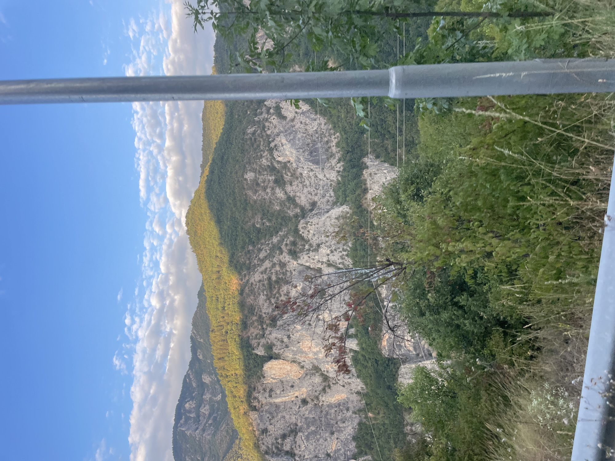 This is a photograph of a mountain landscape with a clear blue sky filled with scattered white clouds. In the foreground, there is a metal pole on the right side of the image. The middle ground is dominated by lush green trees and vegetation, with a prominent rocky cliff face visible behind them. The background features rolling, tree-covered mountains bathed in the warm light of the setting sun, casting shadows across their slopes. The scene appears to be a natural environment, likely in a rural or mountainous region. The image was taken in Yugovo, Bulgaria.
