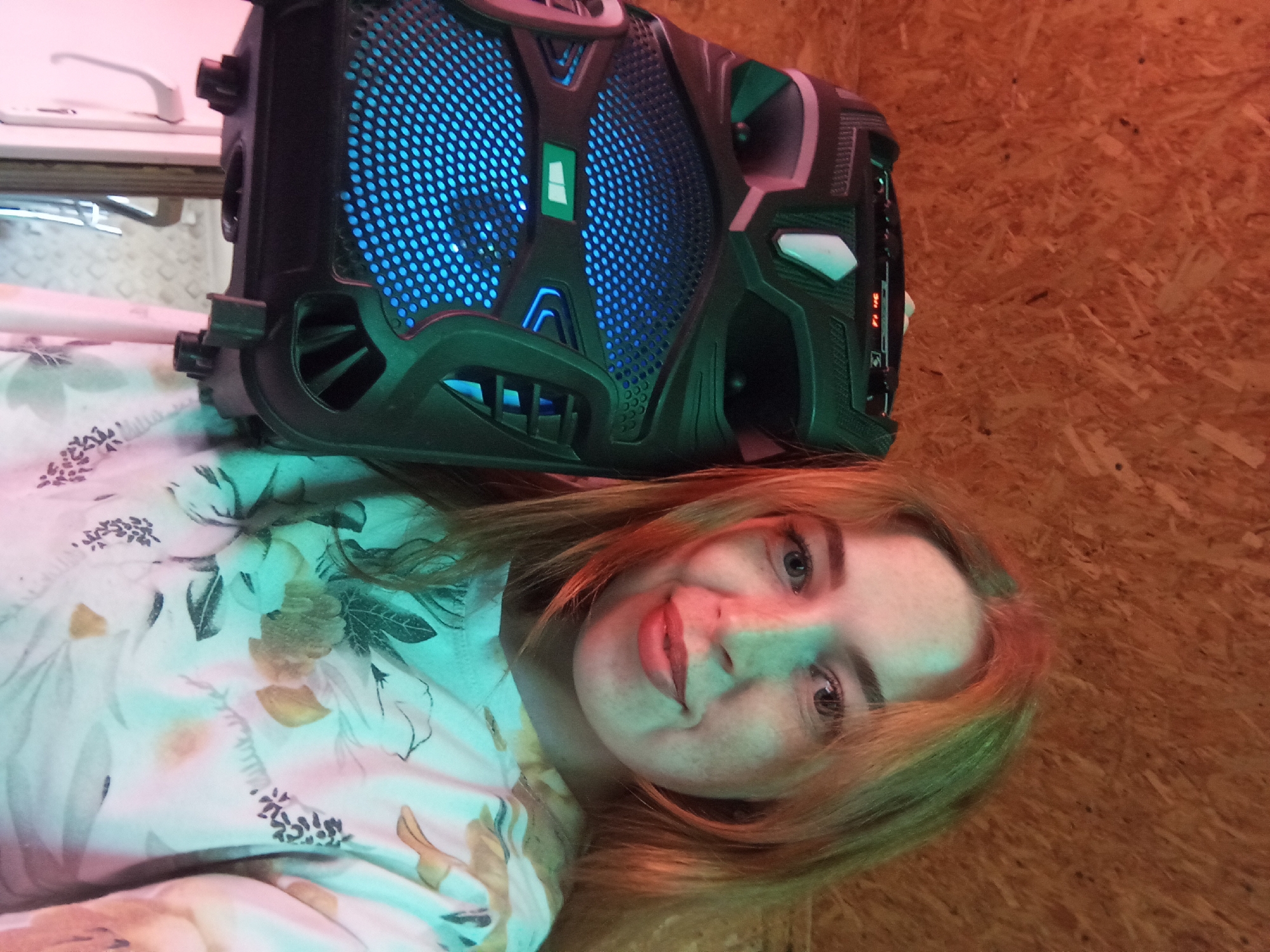 A young fair-skinned person with reddish-blonde hair is lying on what appears to be a floral patterned surface. Resting on or beside the person is a black portable speaker with blue LED lights. The background is a light brown textured material, possibly wood. The city and location are unknown.