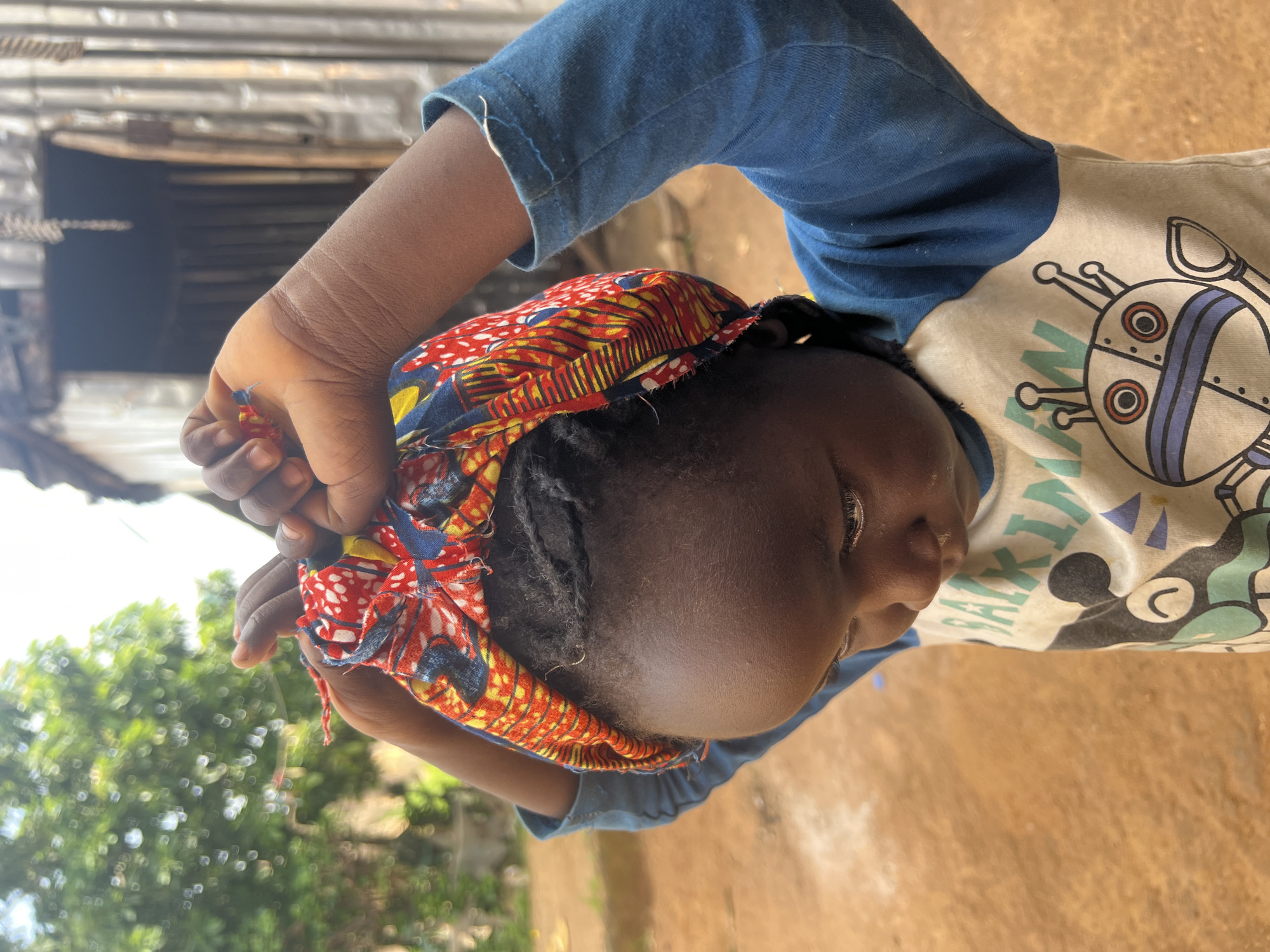 A child with dark skin and braided hair is tying a colorful headscarf around their head. The child is wearing a blue long-sleeved shirt with a graphic design on the front, possibly of a robot. The background appears to be an outdoor setting in a rural or informal urban area, with reddish-brown dirt on the ground and a corrugated metal structure in the background. Green foliage is visible on the left side of the frame. The lighting suggests it is daytime.