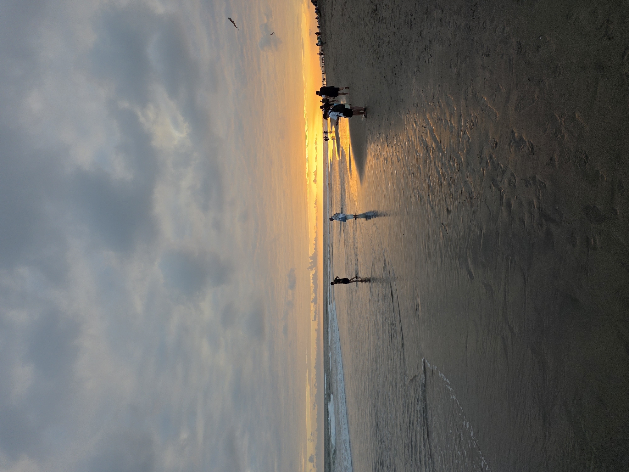 A sunset scene at a beach in North Kuta, Indonesia, shows a group of people walking along the wet sand near the ocean. The sun is low on the horizon, casting a warm orange glow across the sky and reflecting on the water and sand. Waves are gently rolling in from the ocean. The sky is partly cloudy, with some darker gray clouds mixed with lighter, brighter areas where the sun is shining through. The beach itself is sandy, with some footprints visible. Several individuals are scattered across the beach, some walking towards the water and others walking along the shore. The overall atmosphere is calm and serene, characteristic of a beach at sunset.