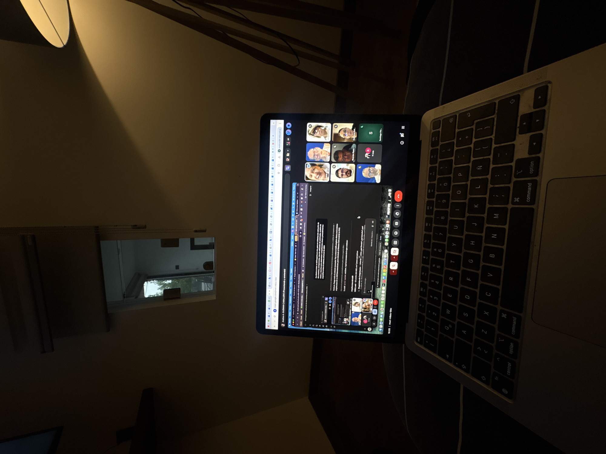 The image shows a laptop with a video conference call in progress, positioned on a dark surface. The background is dimly lit, revealing a lamp and a framed window reflecting a daytime scene. The window's reflection suggests an external environment with greenery, implying an outdoor setting. The video conference shows multiple participants. Based on the OCR text, the call appears to be about a subject called "Institución educativa," and involves participants whose names include "Andres A. Guzman." The call is labeled with a code "mwt-mbau-bbc" and occurred around "18:05". The location of San Bernardino Tlaxcalancingo, Mexico, is not discernible from the image.