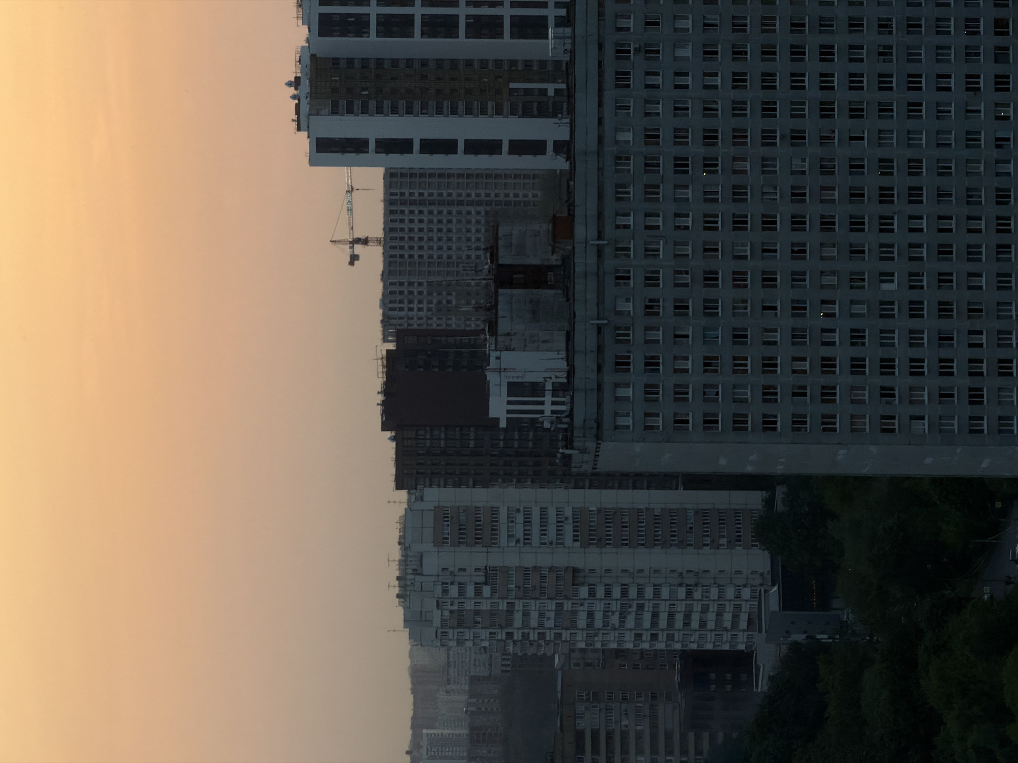 The image shows a cityscape in Khimki, Russia at dusk. The sky is a gradient of light orange to pale yellow. A cluster of tall, gray buildings dominates the scene. Some buildings are under construction, with a crane visible. The buildings have a repetitive grid pattern of windows. Some greenery is visible near the base of the buildings.