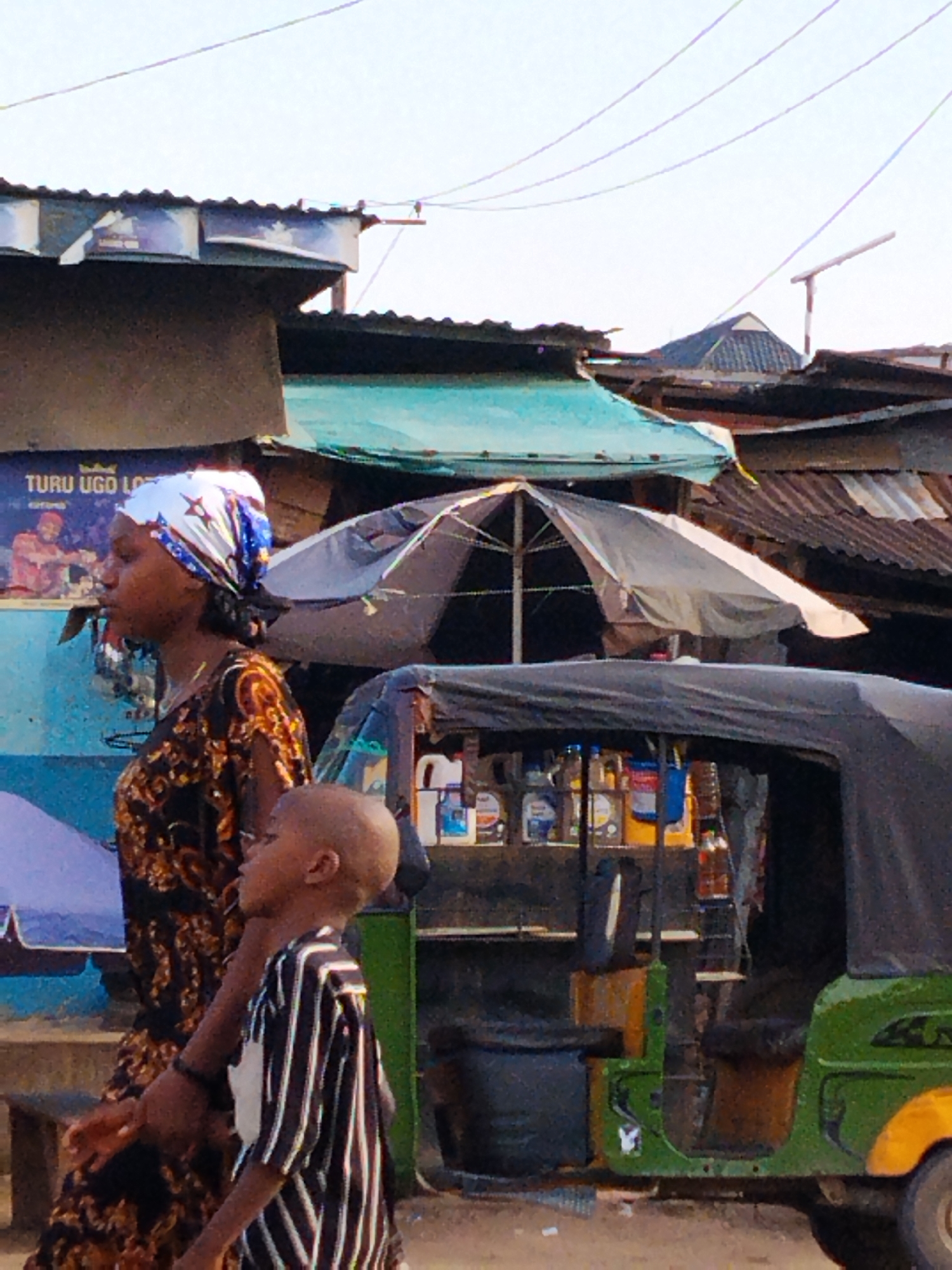The image captures a street scene in Aba, Nigeria. A woman wearing a patterned dress and a headscarf is walking alongside a young boy in a striped shirt. They are on a street with buildings of varying construction, some with corrugated iron roofs. In the background, there is a parked green and black auto rickshaw (tuk-tuk) with items like oil bottles displayed in the vehicle. An advertisement sign is on one of the buildings. Overhead, there are numerous electrical wires stretching across the sky.