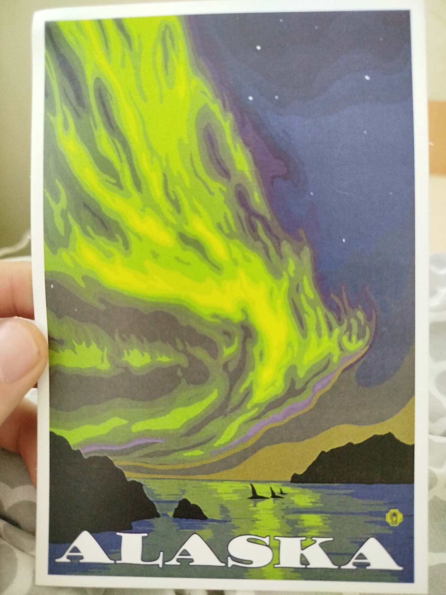 The image is a postcard depicting the aurora borealis over a coastal scene labeled "Alaska". The aurora displays shades of green and yellow streaking across the upper portion of the image. Below the aurora, there's a dark blue and purple sky dotted with stars. The horizon line features landmasses in dark gray and tan, bordering a body of water where the aurora's colors reflect. Three silhouettes, possibly orcas or dolphins, are visible in the water. The word "ALASKA" is printed in large, white block letters along the bottom of the image. There are no elements associated with Varna, Bulgaria.