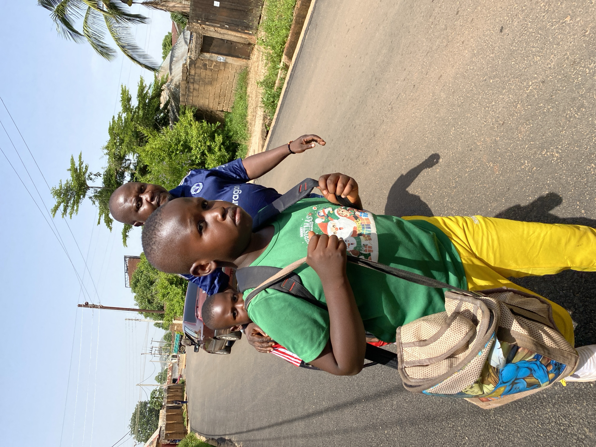 Two young boys and a man are pictured outdoors on a paved road, seemingly on a sunny day, as evidenced by the clear sky and shadows. The man, wearing a blue shirt with a logo, is positioned behind the two boys, with his arm around one of them. The boy in the foreground is wearing a green t-shirt with a Christmas-themed graphic featuring Santa Claus and presents. He also has a backpack with a cartoon print. The other boy, partially visible, is also in a green shirt. Both boys appear to be carrying bags. Yellow pants are visible on one of the individuals, likely one of the boys.
The background shows a street scene with buildings constructed of brick, some with corrugated metal roofs, and lush green trees. Utility poles with numerous wires are also present, indicating an urban or semi-urban environment. A red car is parked on the side of the road. The image is oriented vertically, with the subjects appearing to be walking or standing on the road. The overall impression is one of a casual outdoor moment, possibly during a daytime outing.