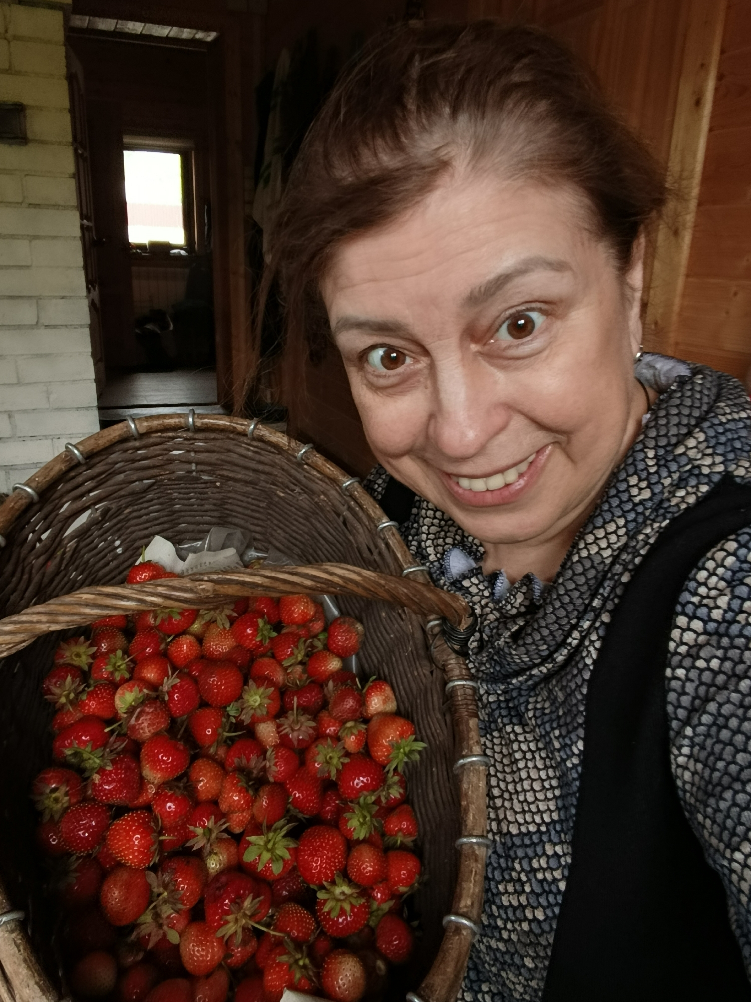 A woman is holding a basket filled with ripe strawberries. She has brown hair and is smiling at the camera. She is wearing a patterned scarf and a black vest. The background appears to be a rustic interior with a wooden wall and a doorway leading to a room with a small window. The scene is likely in a rural or suburban setting, potentially a home in the city of Vereya, Russia, given the context of the image.