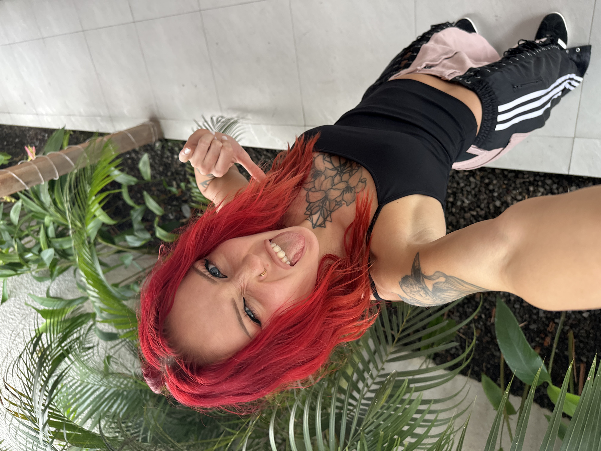 A woman with bright red hair and blue eyes is taking a selfie outdoors. She is wearing a black tank top and pink track pants with white stripes. Her left hand is extended towards the camera, and her right hand is making a fist. She has tattoos on her chest and left arm. The background shows lush green plants and a tiled walkway. The scene is likely in North Kuta, Indonesia, possibly near a resort or a similar establishment given the tropical foliage and tiled pathway.