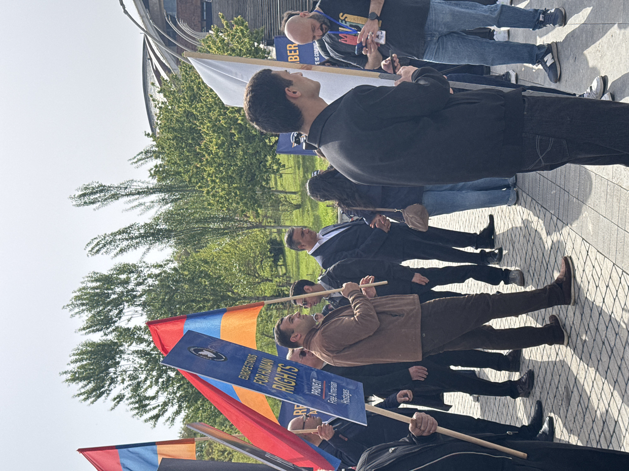 The image shows a group of people in what appears to be a protest or demonstration. They are lined up on a paved area, likely a sidewalk or plaza, with trees and buildings visible in the background. The environment suggests an urban setting, possibly Yerevan, Armenia.
Several people are holding Armenian flags, and a blue sign with white lettering that says "EUROPE STANDS FOR HUMAN RIGHTS" and "PROVE IT Free Armenian Hostages." The people are dressed in casual clothing, including jeans, jackets, and shirts. They are standing and looking towards the right side of the image.