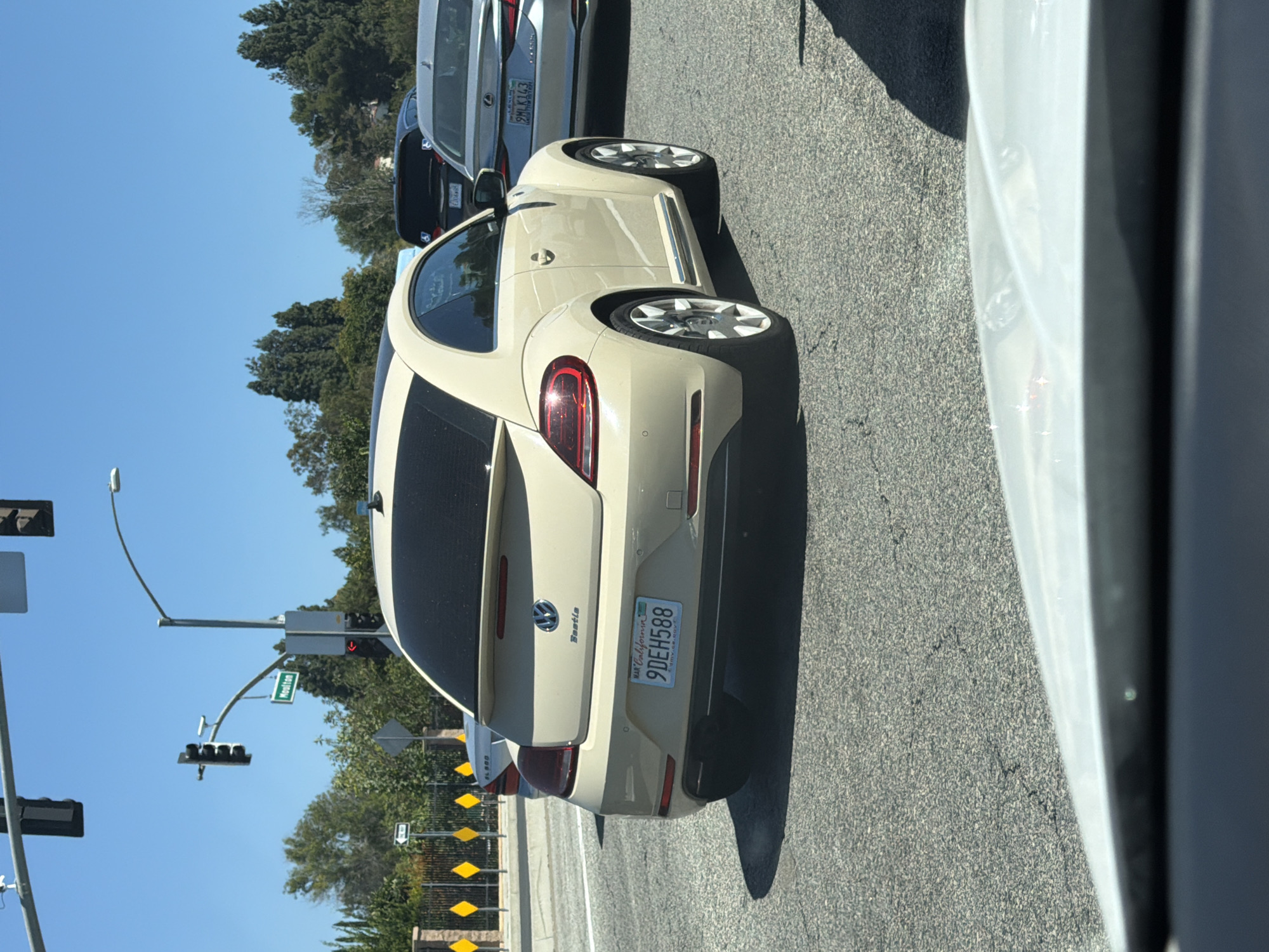 The image captures a street scene in Laguna Woods, California, USA, on a sunny day. The focal point is a light beige Volkswagen Beetle, its rear view prominently displayed. The car bears a California license plate reading "9DEH588". In the background, other vehicles are visible, including a silver Lexus and a dark SUV. Traffic lights and street signs, one indicating "Moulton", are also present, suggesting a traffic intersection. The environment is characterized by asphalt roads, trees, and a clear blue sky. The perspective is from inside another vehicle, with the foreground obscured by the car's dashboard and windshield.
