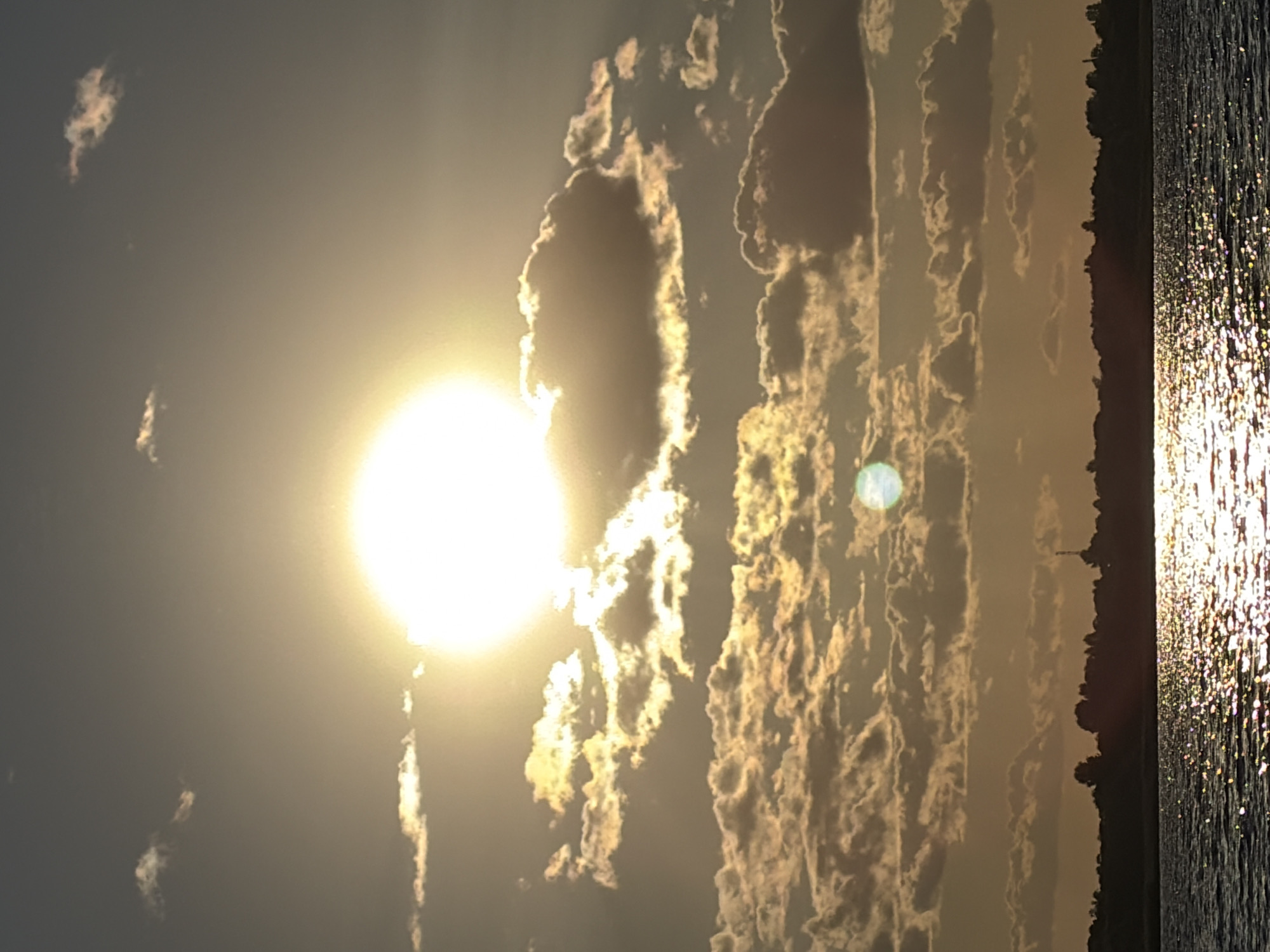 The image depicts a sunset over a body of water, likely a river or lake, in Vidogoshchi, Russia. The sun, a bright, glowing orb, is positioned in the upper center of the frame, partially obscured by scattered clouds that are illuminated by its rays, creating a warm, golden hue. The sky transitions from a bright yellow near the sun to a muted gray towards the top of the image, with a few wispy clouds scattered higher up.
Below the sky is a silhouetted treeline that forms the horizon, suggesting a natural landscape. A faint, distant structure, possibly a tower or antenna, can be seen against the sky within the treeline. The water in the foreground reflects the sunlight, creating a shimmering, dappled effect that stretches across the lower portion of the image. The water appears calm, with gentle ripples visible. There are no people or animals clearly visible in the scene. The overall impression is one of a serene and picturesque natural setting during a vibrant sunset.