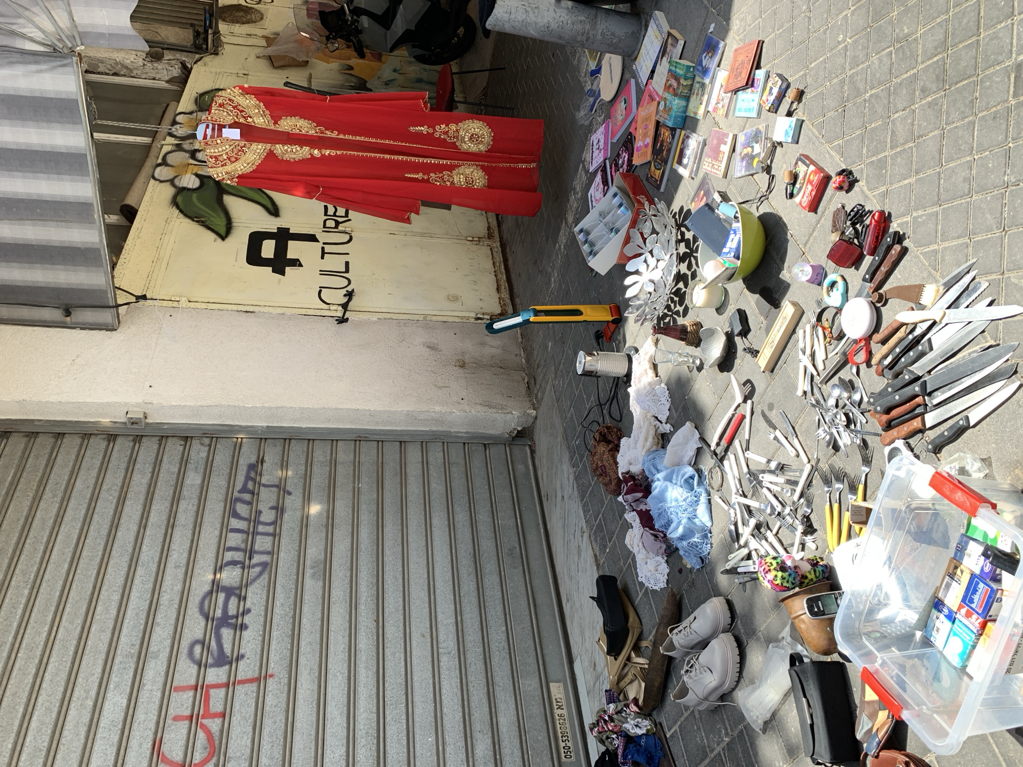 This outdoor scene captures a vibrant street market or yard sale setup in Tel Aviv, Israel, under bright daylight. The ground is paved with grey interlocking pavers. On the left, a light-colored building features a white door with a red garment, possibly a kaftan or traditional dress with intricate gold embroidery, hanging on it. Graffiti on this door includes a stylized black symbol and the word "CULTURE." To the left, a metal roller shutter door is closed, covered in red and purple graffiti, including the number "45" and what appears to be Hebrew script. A small sticker at the bottom of the shutter displays the phone number "050-5390626."
Spread across the paving stones in the foreground and center are numerous miscellaneous items for sale. These include a collection of paperback and hardcover books, with visible titles such as "Wicked" and "I Have to Dream to Live." A wide assortment of kitchen utensils is displayed, including many knives with both white and brown handles, forks, and spoons. Other items include a yellow utility knife, a green bowl, various small decorative objects like a silver floral piece, candles, a Coca-Cola can, and an old Nokia cell phone. Several pieces of crocheted or knitted fabrics in white, light blue, and brown are also laid out. Footwear, including a pair of light grey lace-up shoes and other sandals/heels, is scattered among the goods. A large clear plastic container holds smaller boxes, possibly cigarette packs, and other unidentifiable items. The bright sunshine casts clear shadows, indicating a sunny day, likely morning or early afternoon. No people are visible in the frame.