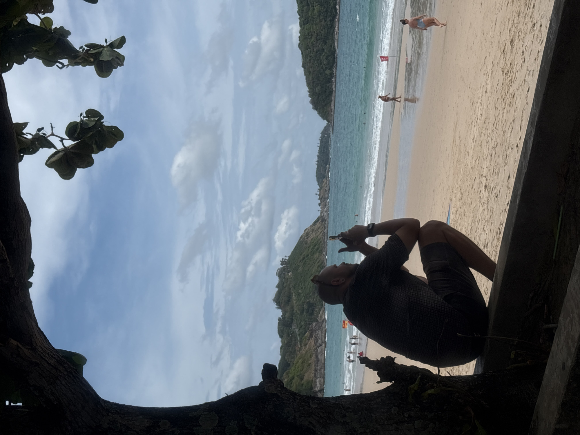 The image shows a view of a beach in Rawai, Thailand, framed by foreground elements including a dark, shadowed tree branch with leaves and a section of a dark wooden structure. A person is positioned on a branch or ledge, appearing in silhouette, with their legs drawn up and arms raised toward their face. The beach is light-colored sand, and the water is a turquoise color with waves breaking near the shore. Several people are visible on the beach and in the water. A land mass covered in trees is also seen extending into the water. The sky is blue with scattered clouds.