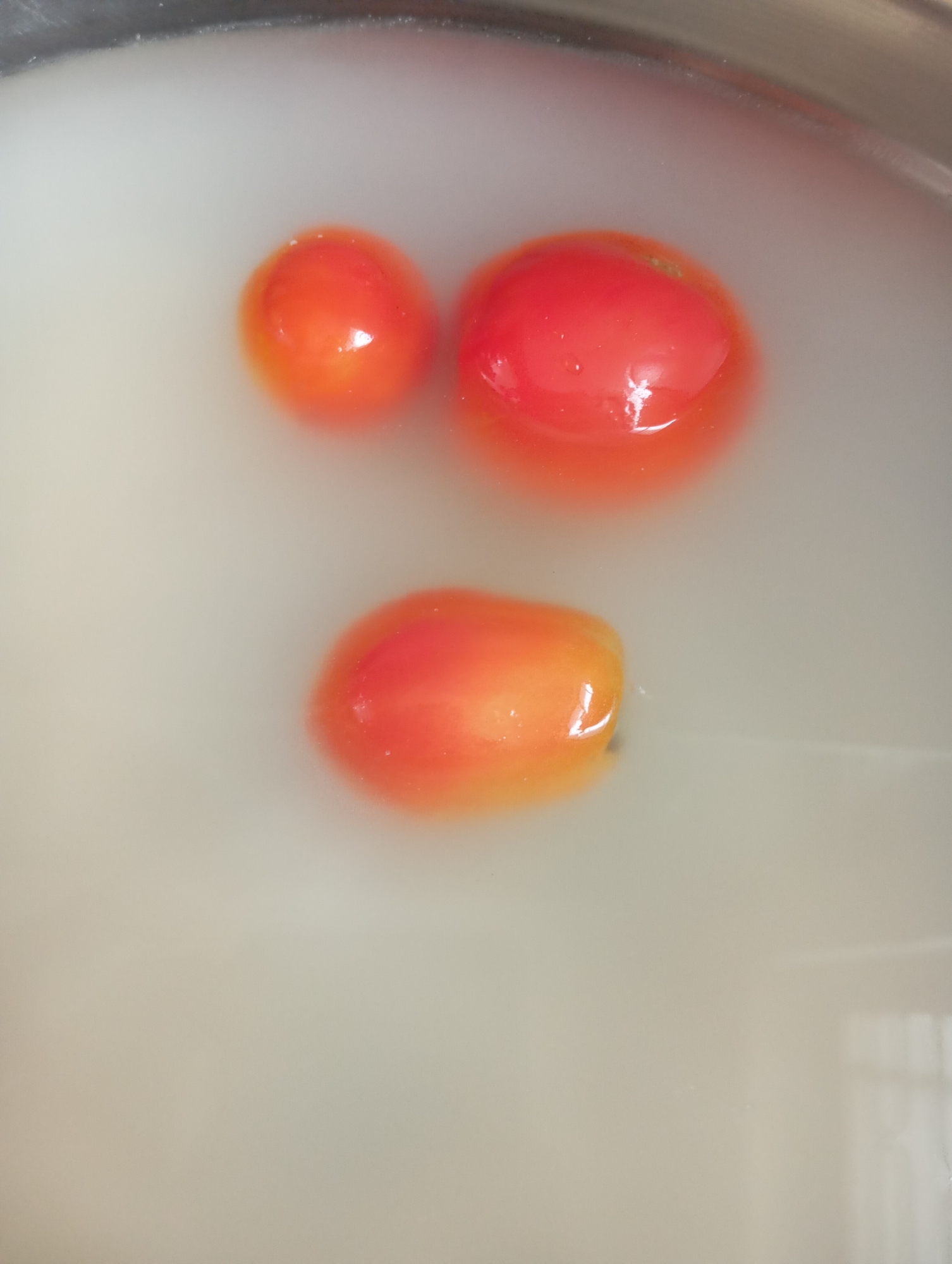 Three small, ripe tomatoes are submerged in a cloudy liquid, likely water or milk, within a metallic basin. The tomatoes, varying slightly in size and shape, exhibit shades of red and orange, with some reflecting highlights on their smooth surfaces. The liquid itself is opaque and creamy, obscuring the bottom of the basin. The overall impression is a close-up shot of produce being prepared or cleaned. The location is Unknown, Unknown.