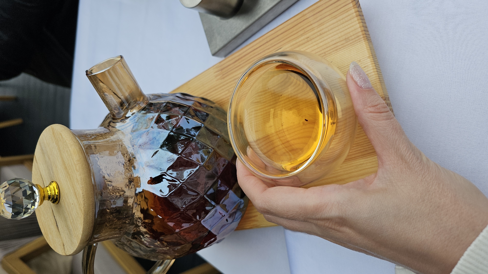 An overhead shot shows a hand holding a clear glass teacup filled with amber-colored liquid. The teacup rests on a light-colored wooden board, which is placed on a white tablecloth. Behind the teacup is a faceted glass teapot with a wooden lid topped by a crystal finial. The teapot contains a dark amber liquid. The scene is set indoors, likely in a cafe or restaurant in Minsk, Belarus. The lighting is natural, creating reflections on the glass surfaces. The background is blurred, but appears to be an indoor setting with some indistinct furniture.