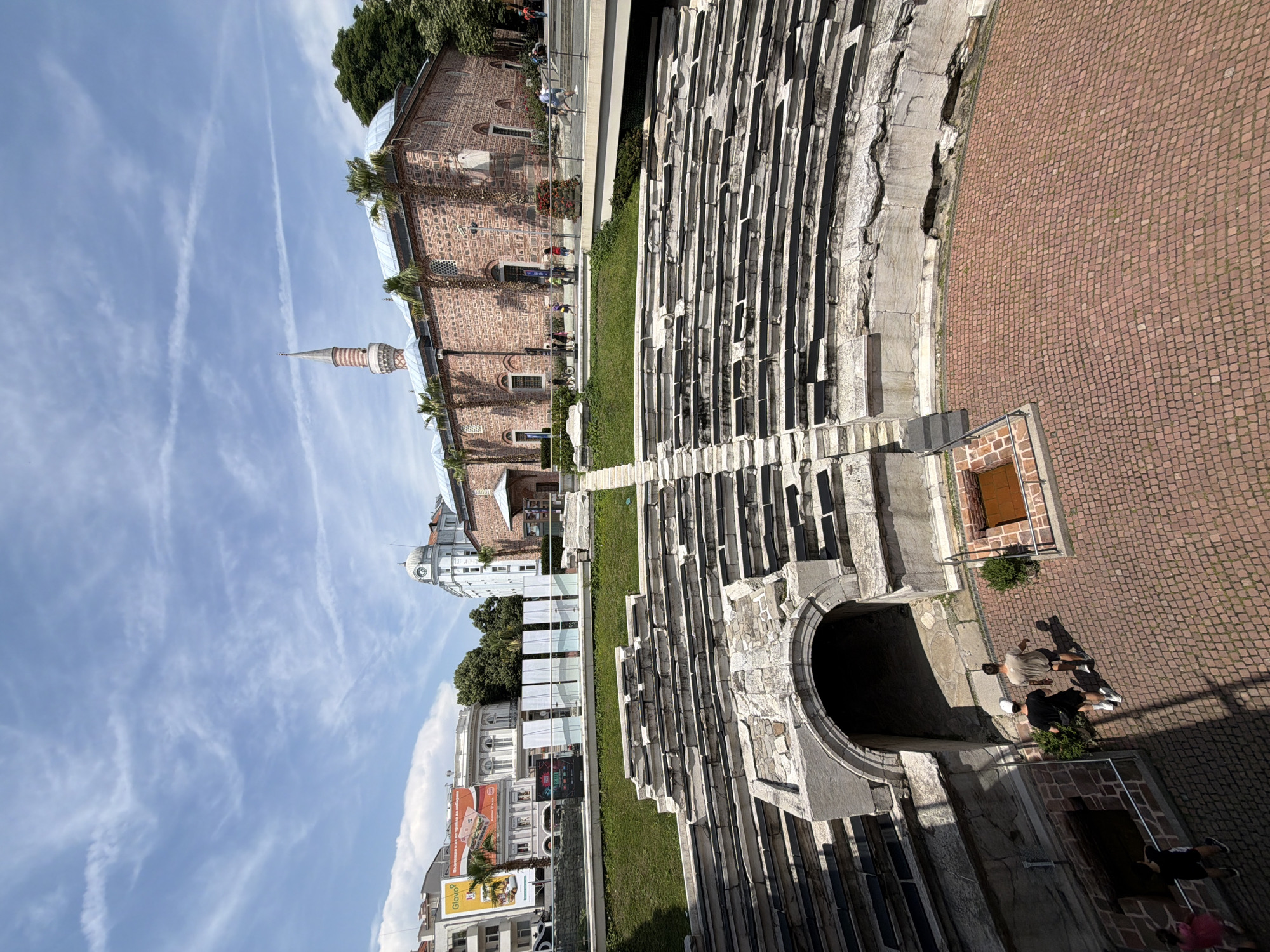 This image captures a wide-angle view of the Roman Stadium of Plovdiv, Bulgaria, with the Dzhumaya Mosque in the background, under a bright, partly cloudy sky. The foreground features the well-preserved lower tiers of the ancient stadium's seating, constructed from stone. An arched, dark entrance/passage is visible on the lower left, leading onto the stadium's arena floor, which is paved with reddish-brown cobblestones. Two men, wearing a white cap and a black t-shirt, and a beige t-shirt respectively, walk towards the right on this paved area. Two additional figures are partially visible to the far left.
Above the stadium seating, a sloped grassy area leads to a raised pedestrian walkway or bridge. Several individuals are observed walking along this elevated path. Beyond the walkway, the prominent, red-brick Dzhumaya Mosque stands with its distinctive striped minaret rising on the left side of its roofline. Palm trees are visible along the mosque's facade. To the left of the mosque and behind the stadium, various modern urban buildings are present, including a light-colored building with a domed clock tower. A large billboard displaying "Glovo" and Cyrillic text is visible above these buildings. The sky is bright blue with scattered white, wispy clouds and visible contrails. The strong sunlight casts distinct shadows across the scene.