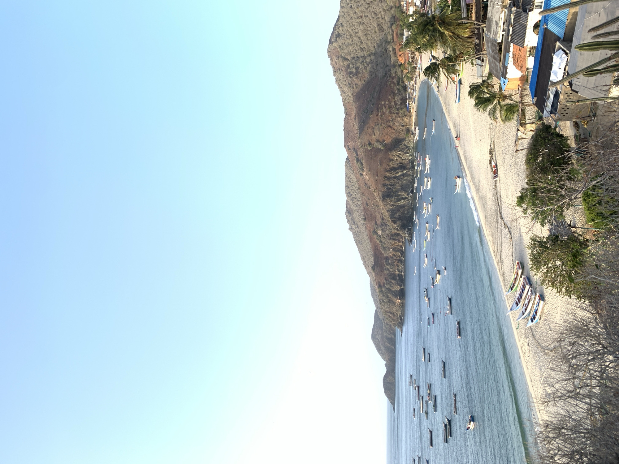 This image captures a serene coastal landscape in Pedro González, Venezuela, featuring a tranquil bay under a bright, clear blue sky. The scene is dominated by a wide expanse of light-colored sandy beach that gently curves along the edge of calm, clear blue water.
Numerous small, long, narrow boats, likely fishing vessels or canoes, are dispersed across the bay's surface, indicating local maritime activity. Further down the beach, several more colorful boats are pulled ashore, resting on the sand. The coastline is backed by an impressive range of hills or mountains, covered in dry, sparse vegetation, typical of certain arid coastal regions.
Along the upper edge of the beach, a small collection of buildings forms a settlement. These structures appear modest, with some featuring distinctive blue corrugated metal roofs, nestled amidst green palm trees. In the immediate foreground on the right, drier, bare trees and cacti are visible.
The bright sunlight and unblemished sky suggest a clear day, likely late morning or early afternoon. No individuals are explicitly visible, but the presence of boats and structures implies an active, peaceful coastal community. The overall mood is calm and picturesque. There is no visible text in the image.